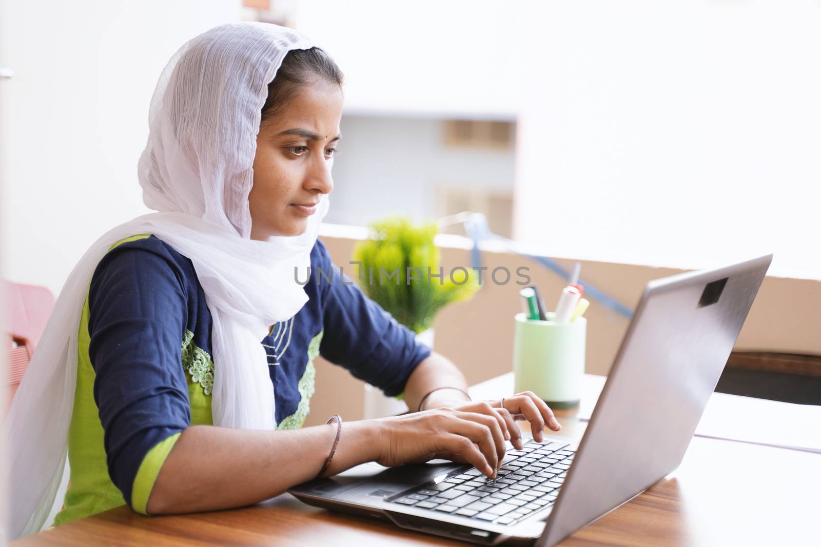 Happy young indian girl using computer looking at screen working in internet - young girl busy working or reading on laptop at home - concept of online education, work from home or wfh by lakshmiprasad.maski@gmai.com