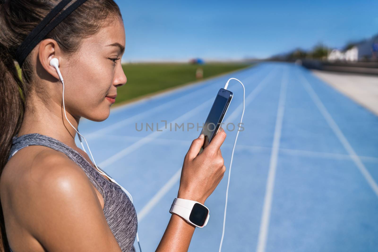 Running music motivation woman listening to phone app with headphones. Runner looking at smartphone on stadium running track with earphones and mobile phone ready to run. Healthy lifestyle by Maridav