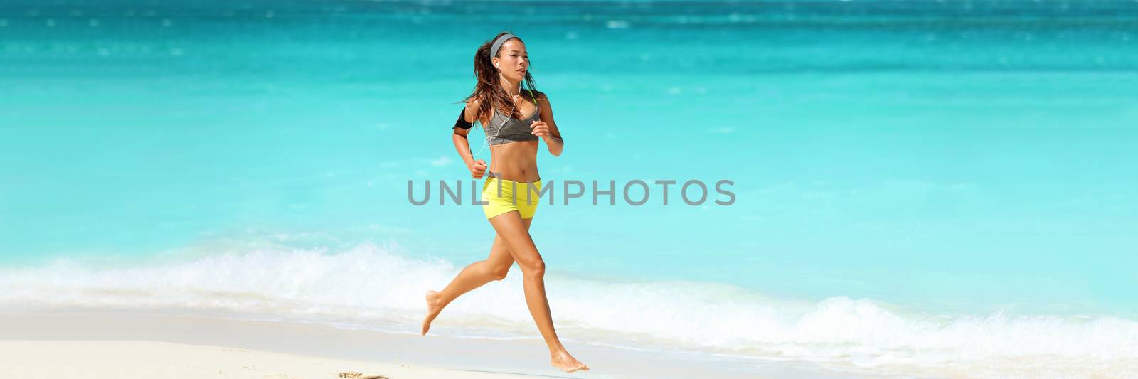 Healthy active lifestyle banner panorama. Fitness woman working out cardio running on beach wearing armband and earphones listening to phone music training jogging with blue background copy space.