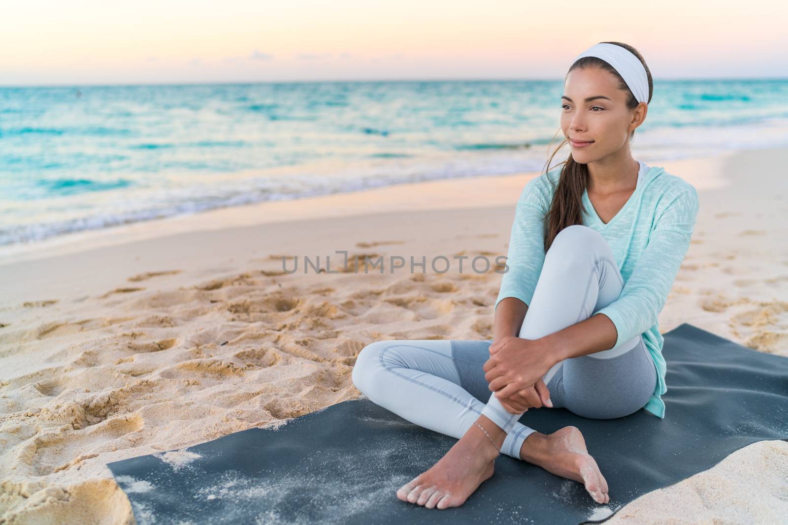 Yoga beach woman relaxing on fitness mat after workout. Fit Asian fitness athlete girl doing exercise in activewear at sunset or sunrise. Mindfulness healthy lifestyle.