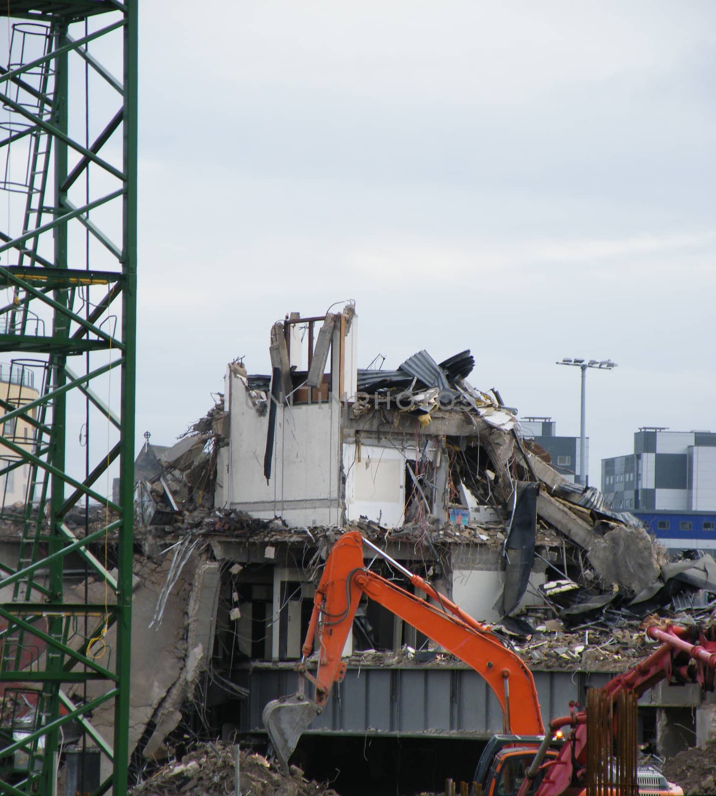 an orange digger with a backhoe working on a large urban demolition site with a metal tower crane at the edge of the frame and grey sky