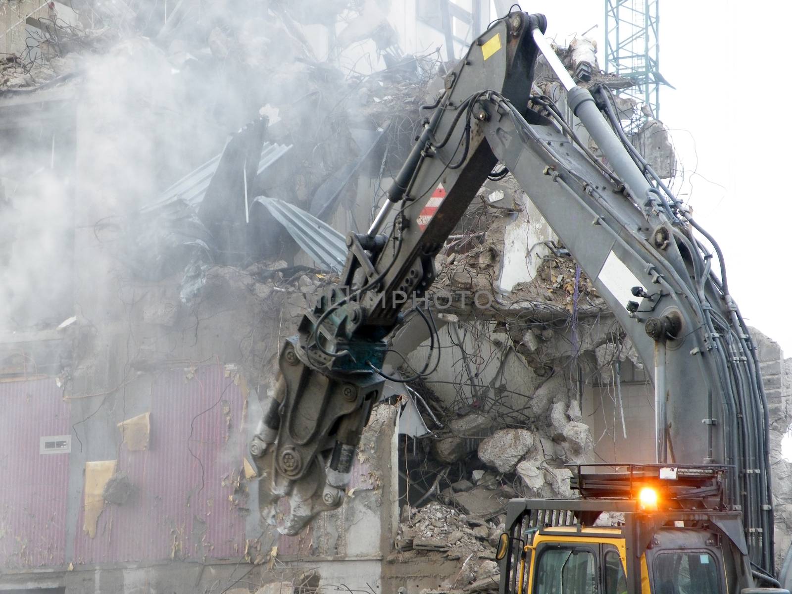 A mechanical digger with a pneumatic hammer tearing down and old building on a an urban development site with dust debris and wires visible