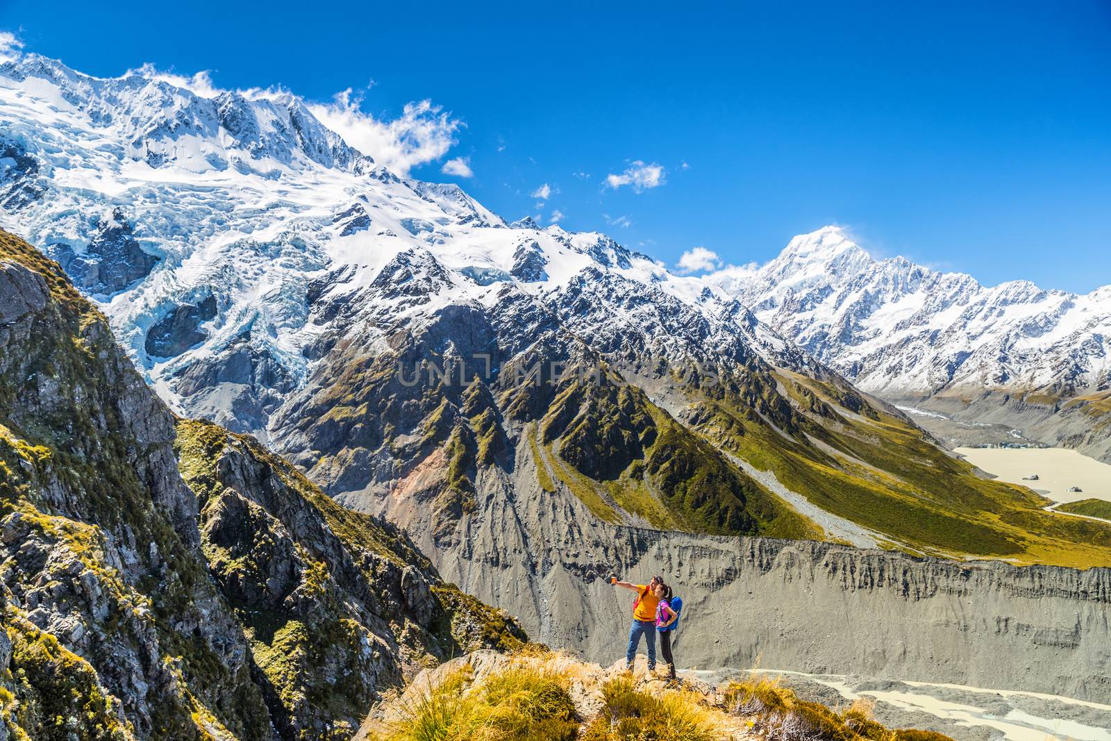 Backpackers couple hiking taking selfie phone picture at Mount Cook mountains view. People tramping in New Zealand. People hikers taking self-portrait photos during hike in alps of south island.