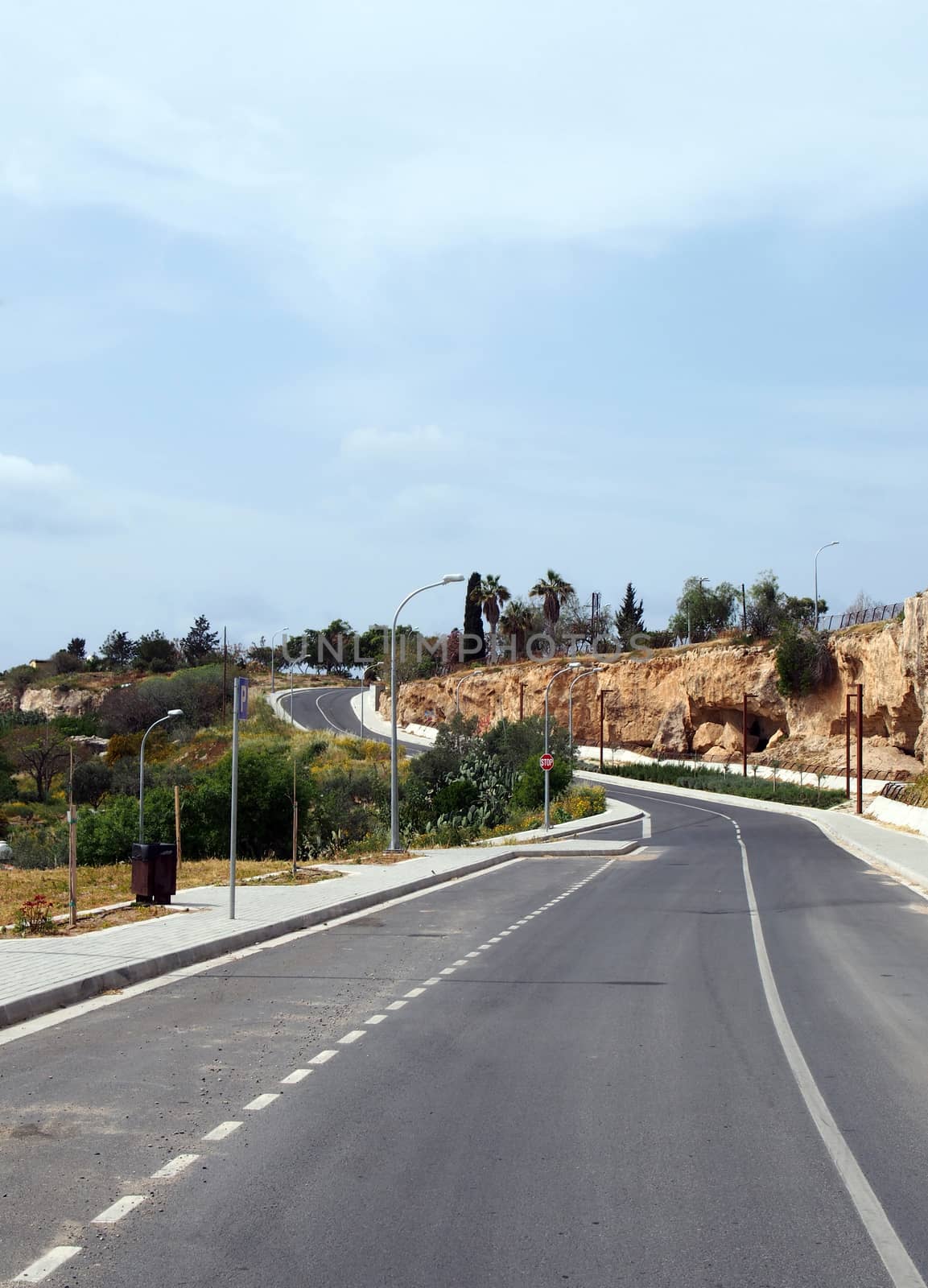 an empty two lame road with pavements and lampposts curving up a hill into the distance with surrounding trees and blue sunlit sky