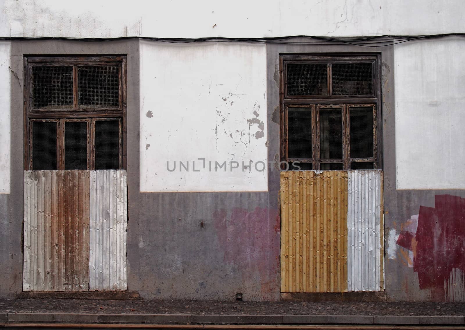 the front of a white and grey painted abandoned house with broken doors and windows boarded up with rusty corrugated iron with splashed of red and yellow