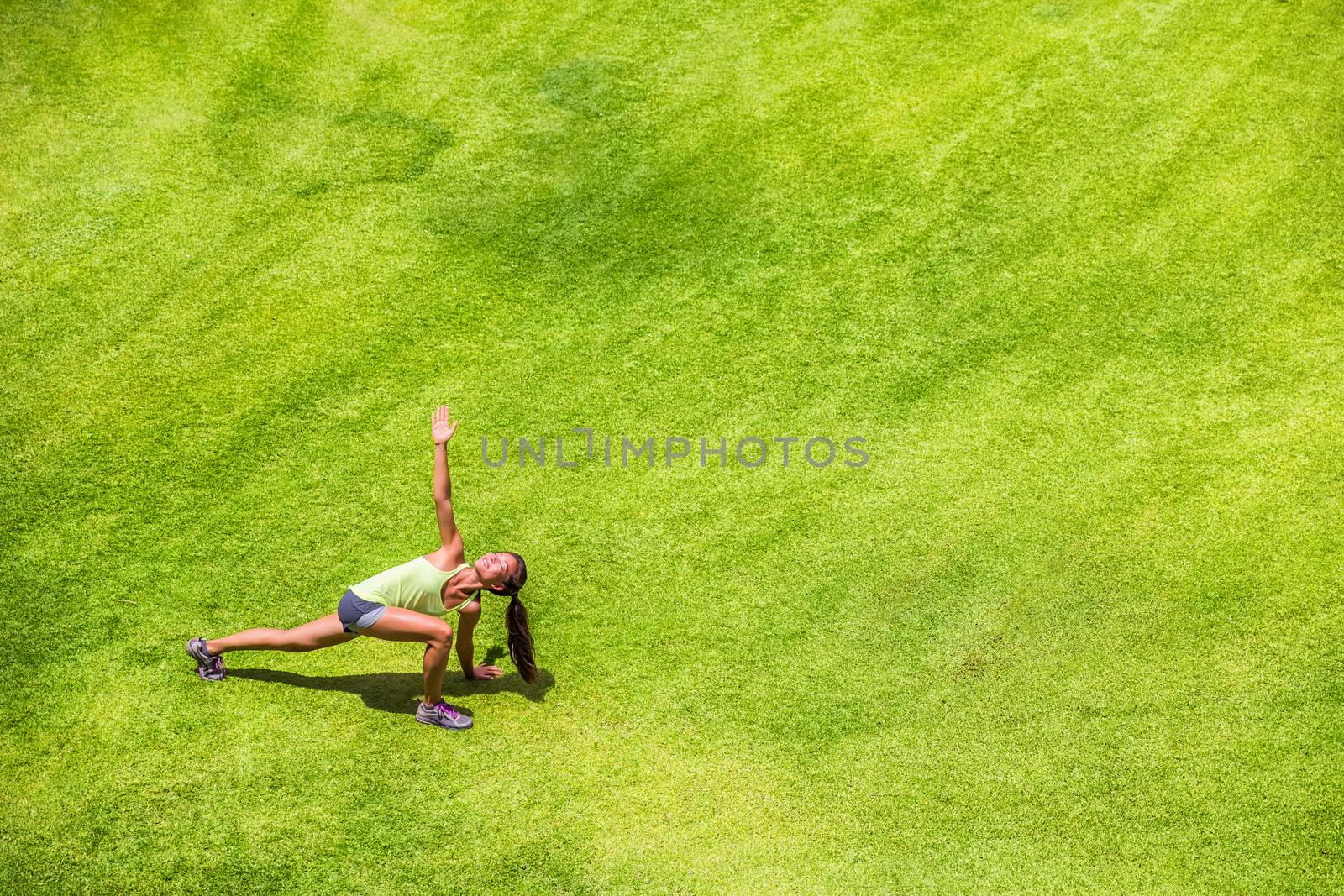 Runner woman stretching legs running warm-up doing twist lunge on green grass park background. Happy jogging girl training on summer outdoors by Maridav