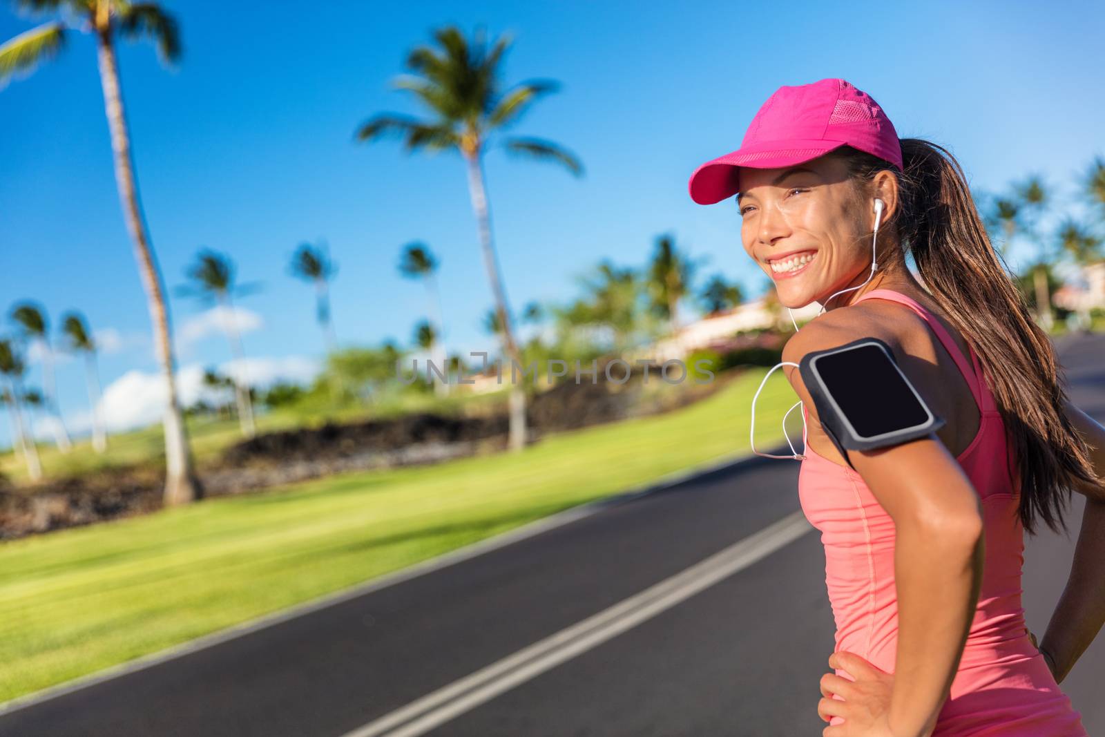 Fitness motivation running music woman runner. Happy jogging girl listening to smartphone app with earphones. Asian athlete wearing cap and armband smart phone holder outdoor on city road by Maridav