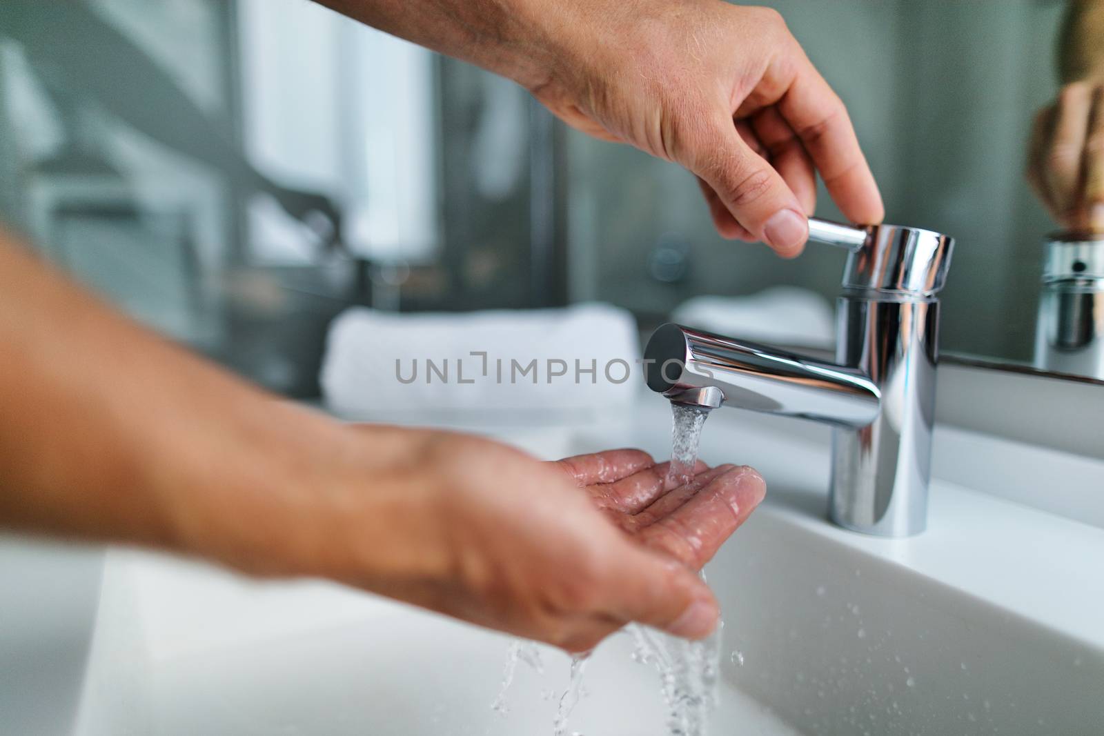 Man washing hands in bathroom sink at home checking temperature touching running water with hand. Closeup on fingers under hot water out of a faucet of a sink by Maridav