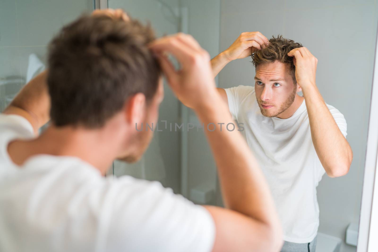 Hair loss man looking in bathroom mirror putting wax touching his hair styling or checking for hair loss problem. Male problem of losing hairs by Maridav