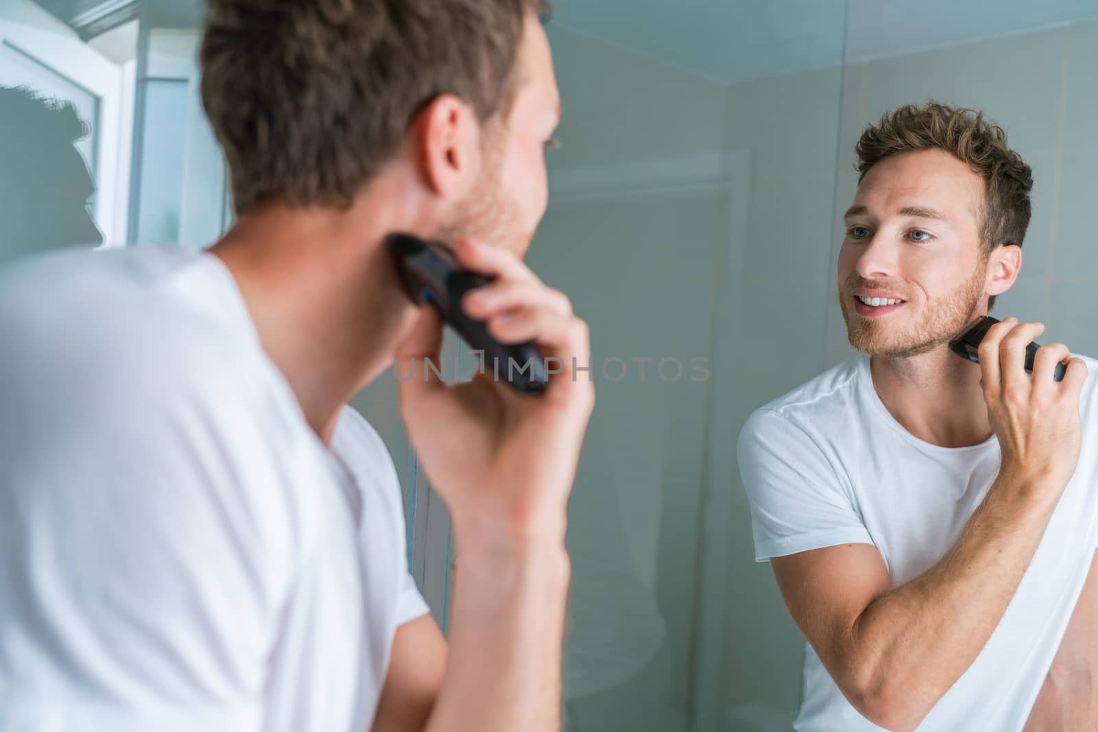 Shaving beard with electric shaver in bathroom. Man looking in mirror, male beauty care routine lifestyle by Maridav