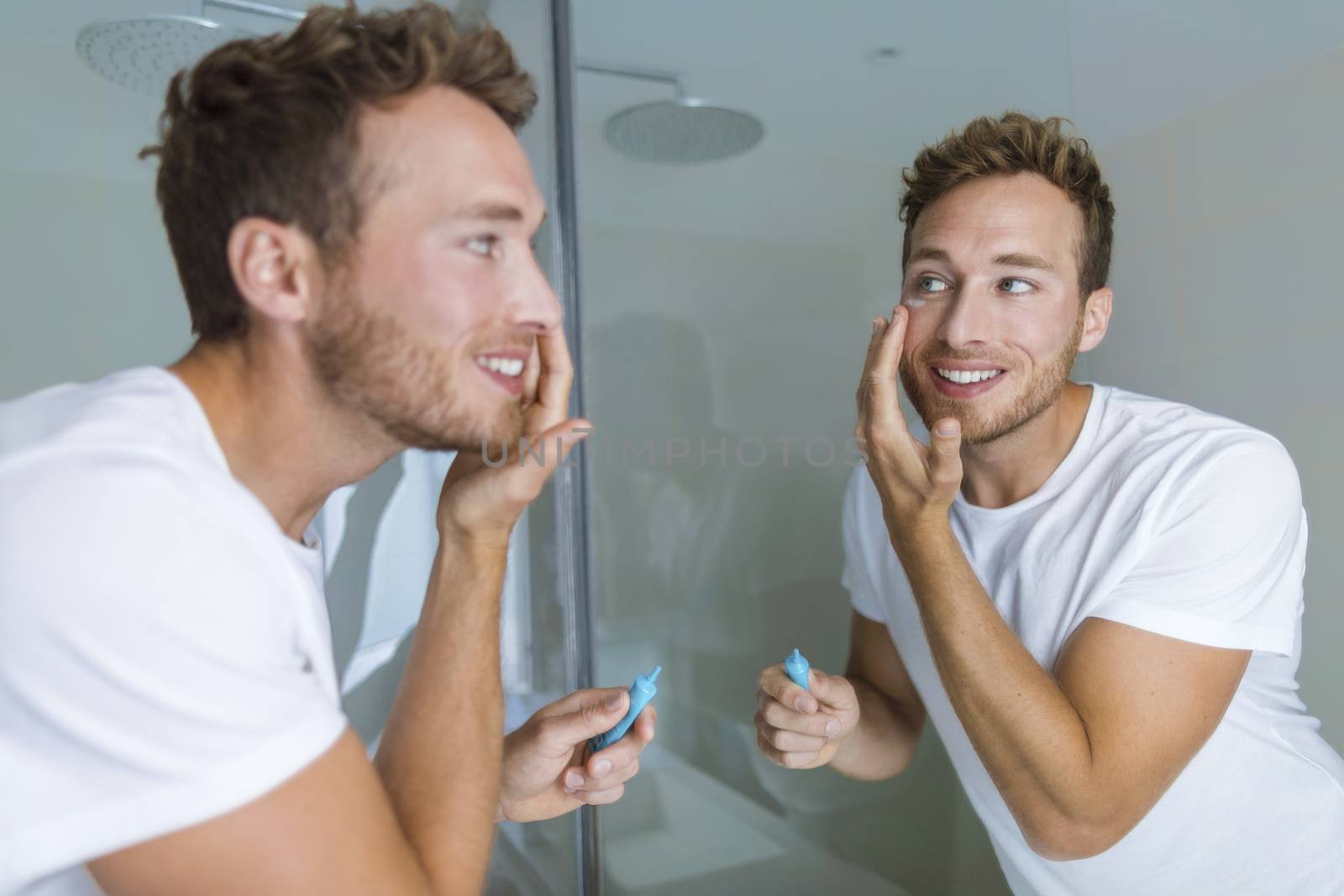 Man putting skincare facial treatment cream on face. Anti-aging skin care product. Male beauty morning routine at home lifestyle. Guy looking in bathroom mirror applying moisturizer under eyes.