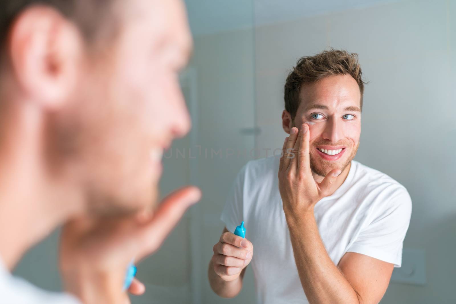 Smiling young man putting face cream under eyes to treat wrinkles or undereye eyebags, Anti-aging facial treatment. Male beauty by Maridav