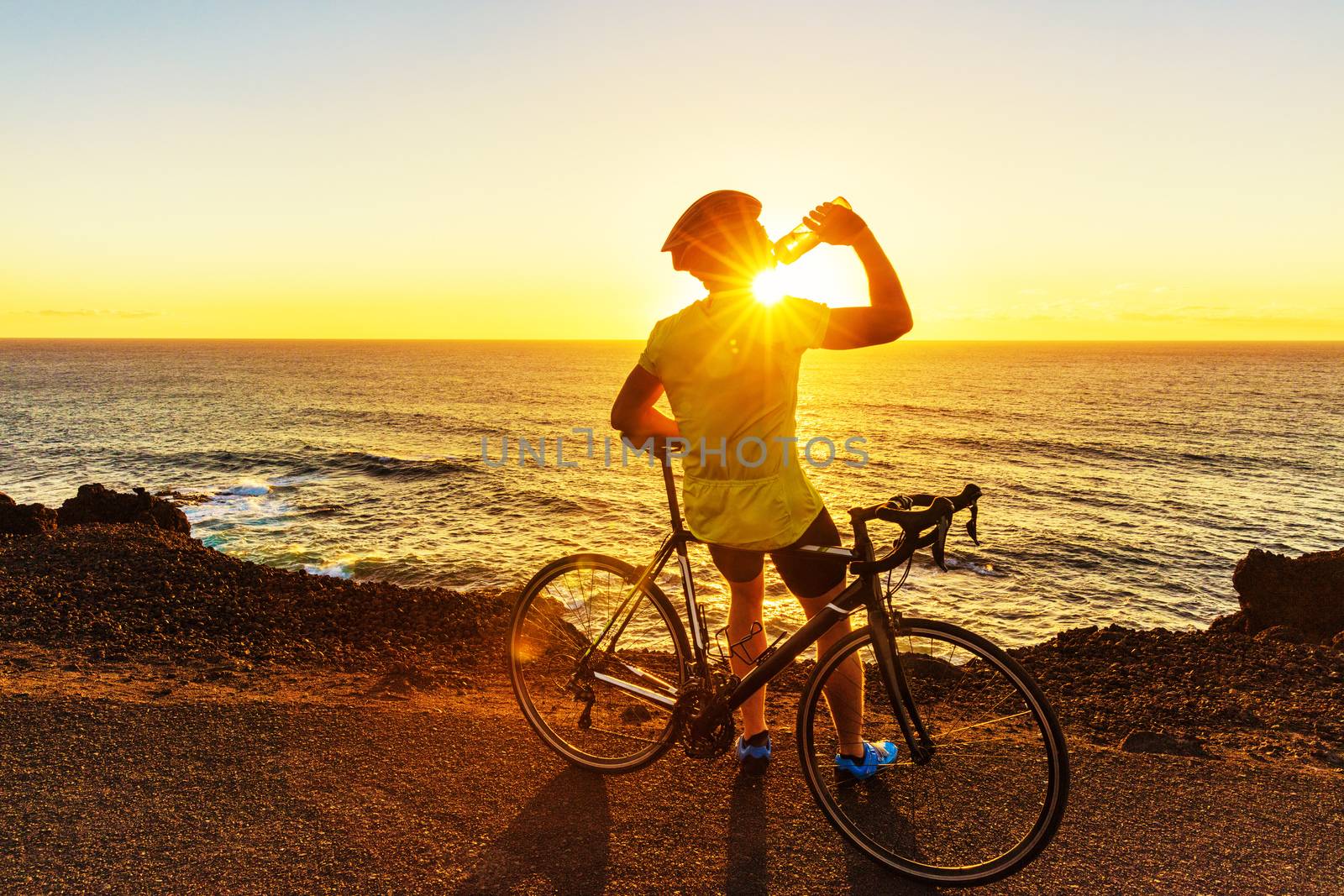 Athlete cyclist man drinking water after intensive biking training, enjoying sunset and ocean. Healthy active lifestyle sports fitness man resting on bike with sun flare.