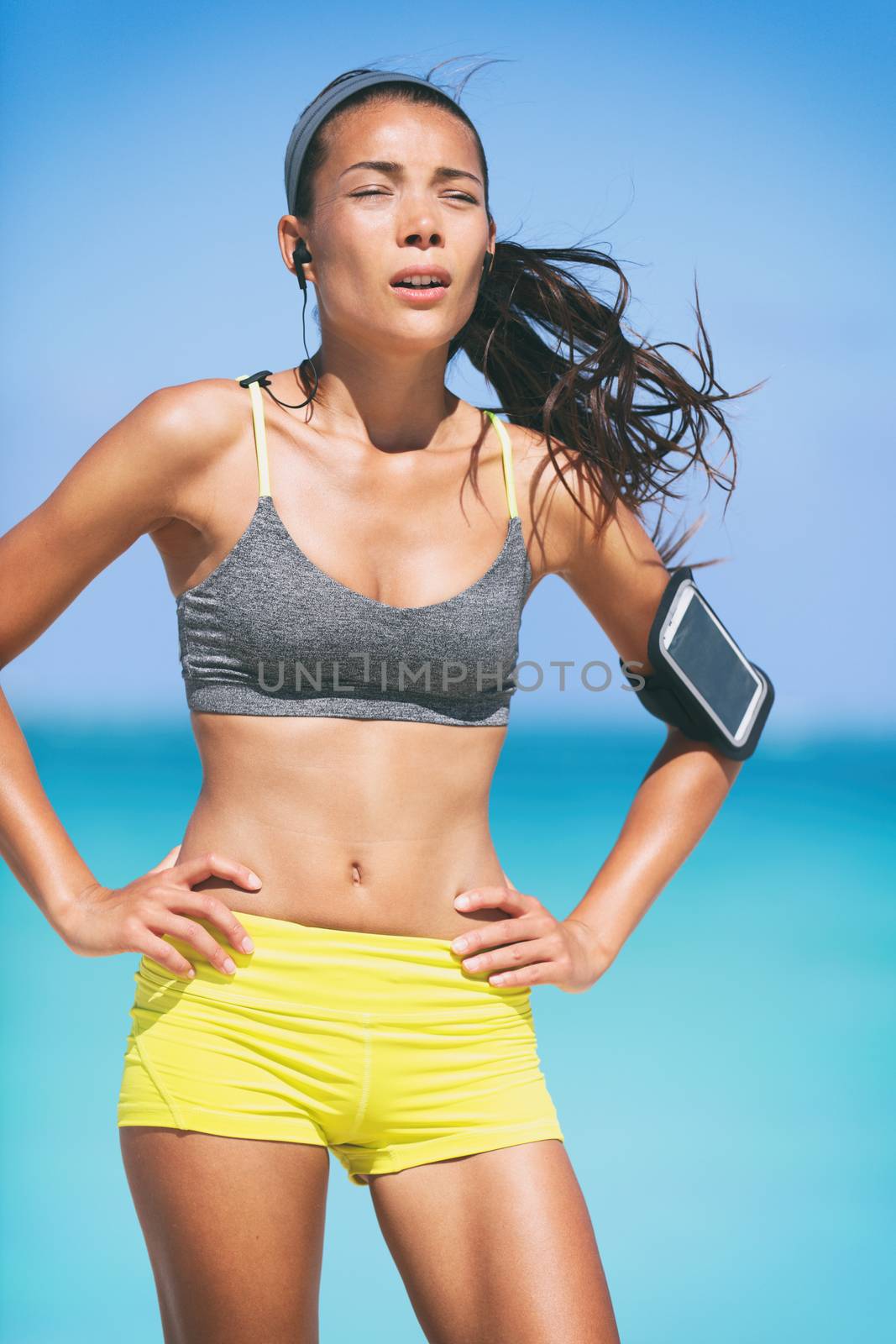Sport fitness runner woman tired breathing during difficult workout. Heat exhaustion dehydrated jogging girl sweating listening to earphones music with mobile phone armband by Maridav