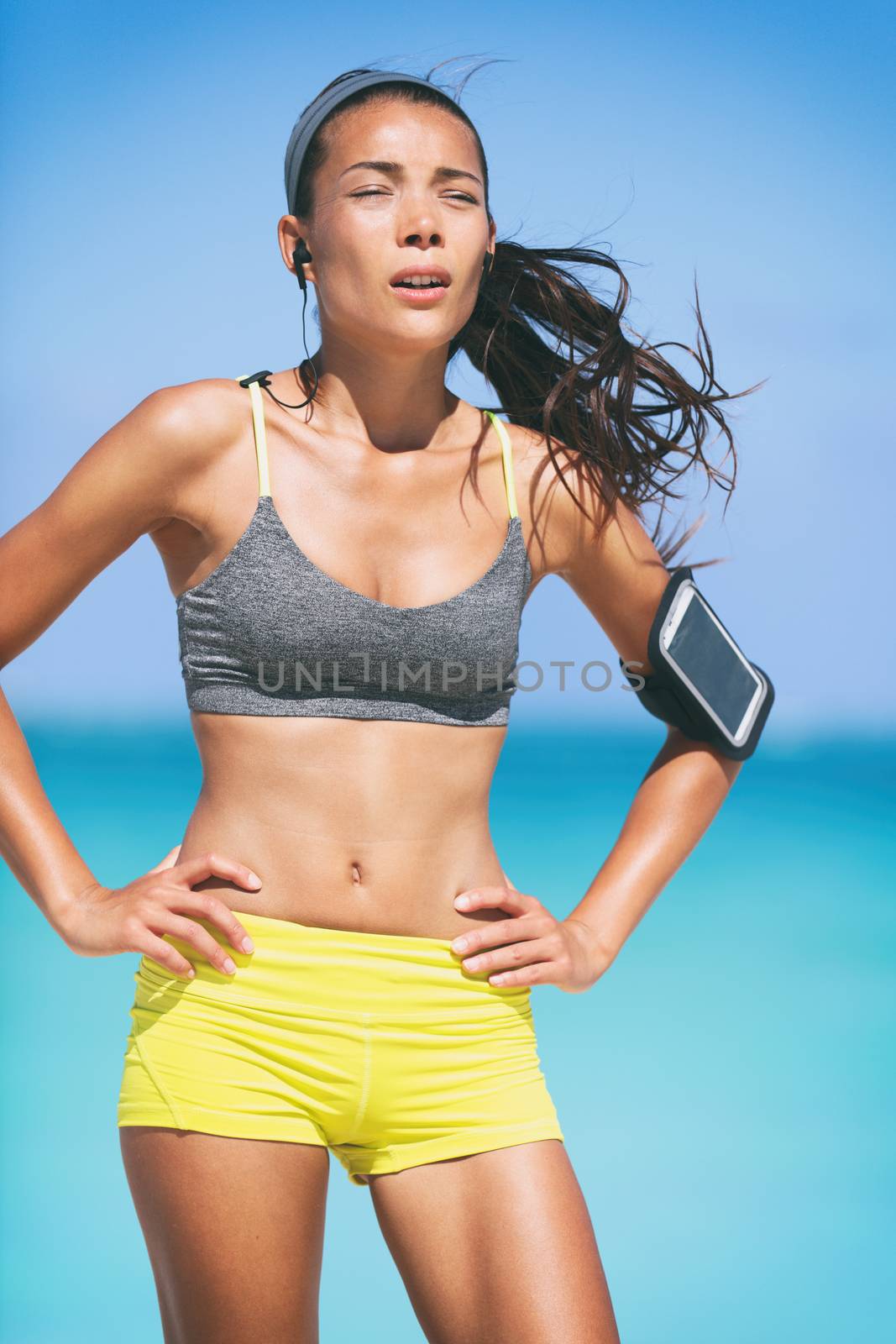 Sport fitness runner woman tired breathing during difficult workout. Heat exhaustion dehydrated jogging girl sweating listening to earphones music with mobile phone armband.