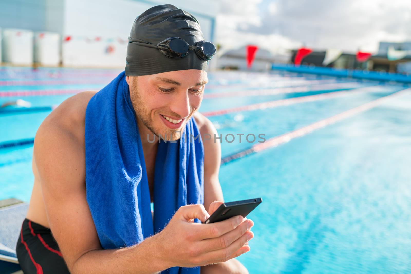 Swimmer man with swimming cap and goggles at pool using his mobile phone texting on smartphone app after training in outdoor pool. Happy athlete holding cellphone at fitness centre.