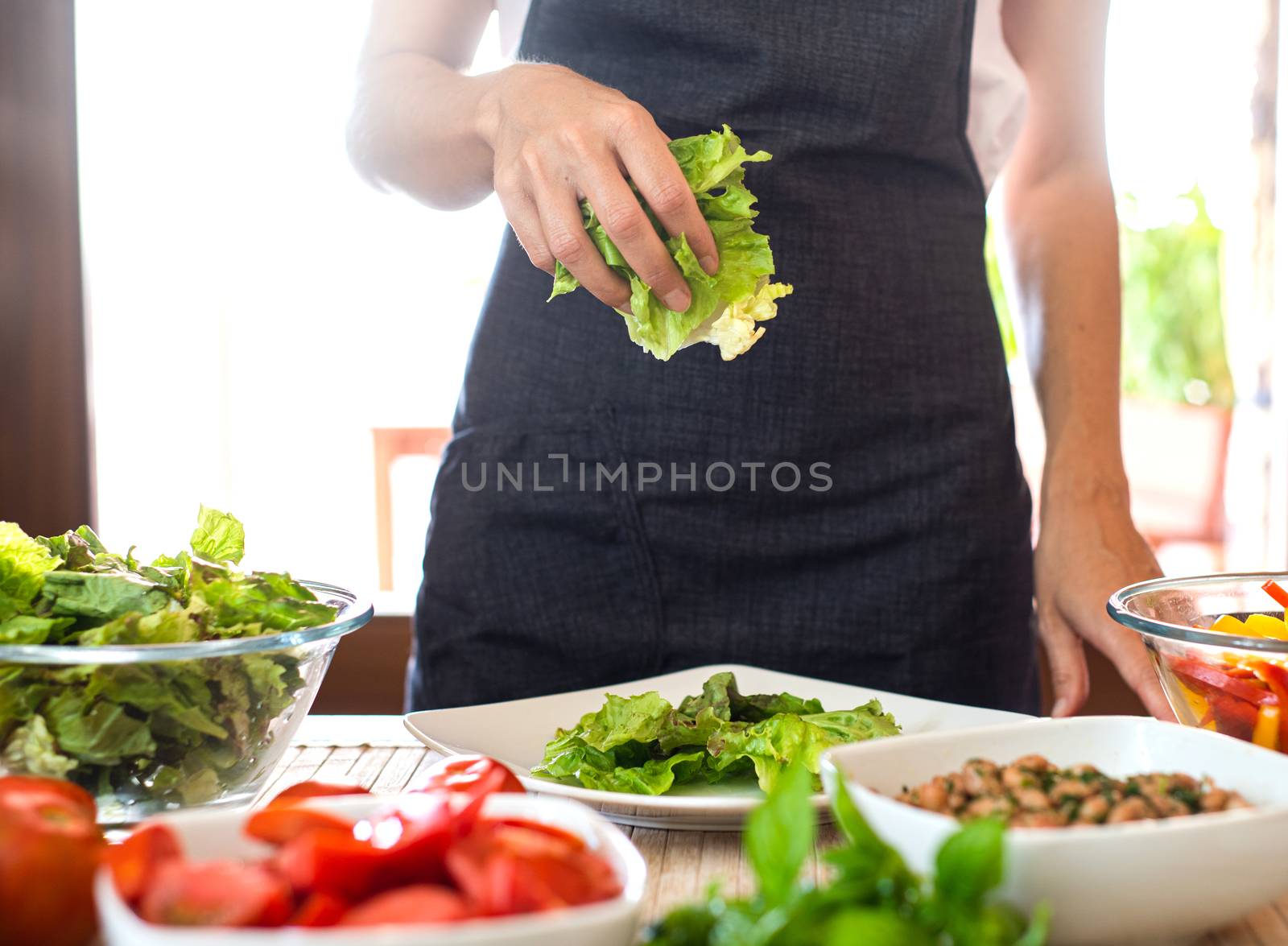 Woman chef preparing a fresh salad at home with sunlight in background with lettuce, tomatoes, beans, seeds on the table - Healthy vegan and vegetarian food concept by robbyfontanesi