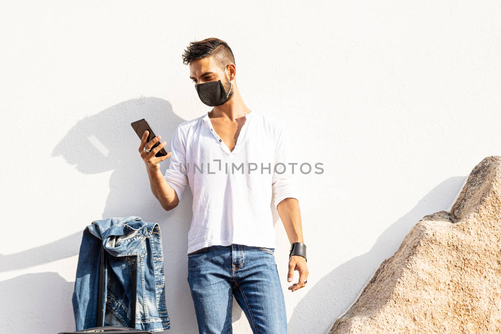 Stylish millennial boy standing to a white wall with COVID protection mask using his smartphone - Solo traveller safe concept - Connected young man communicating with technology by robbyfontanesi