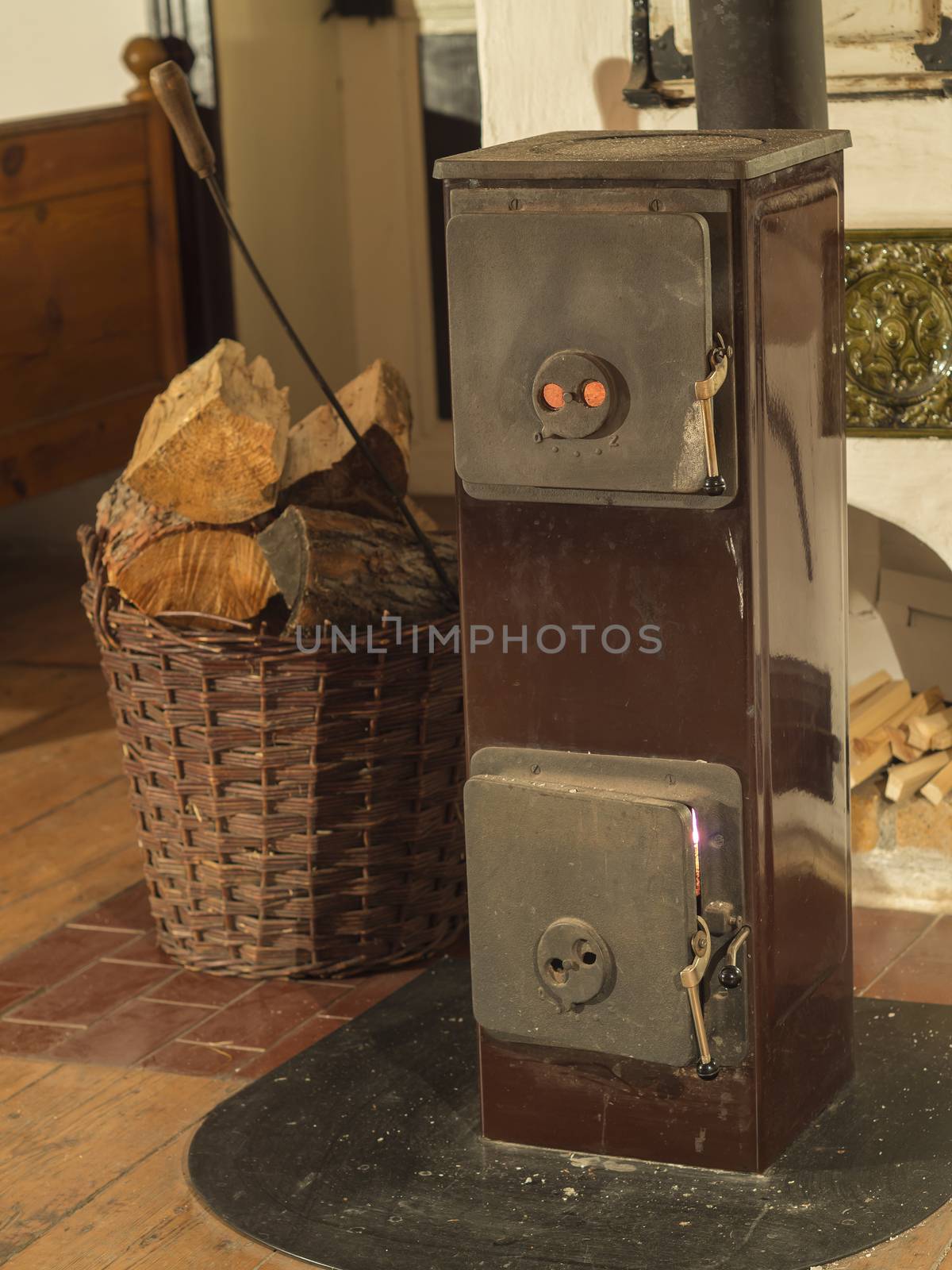 old metal burning stove with wicker basket full of chopped wood and fire-hook on wooden board floor in timbered cottage
