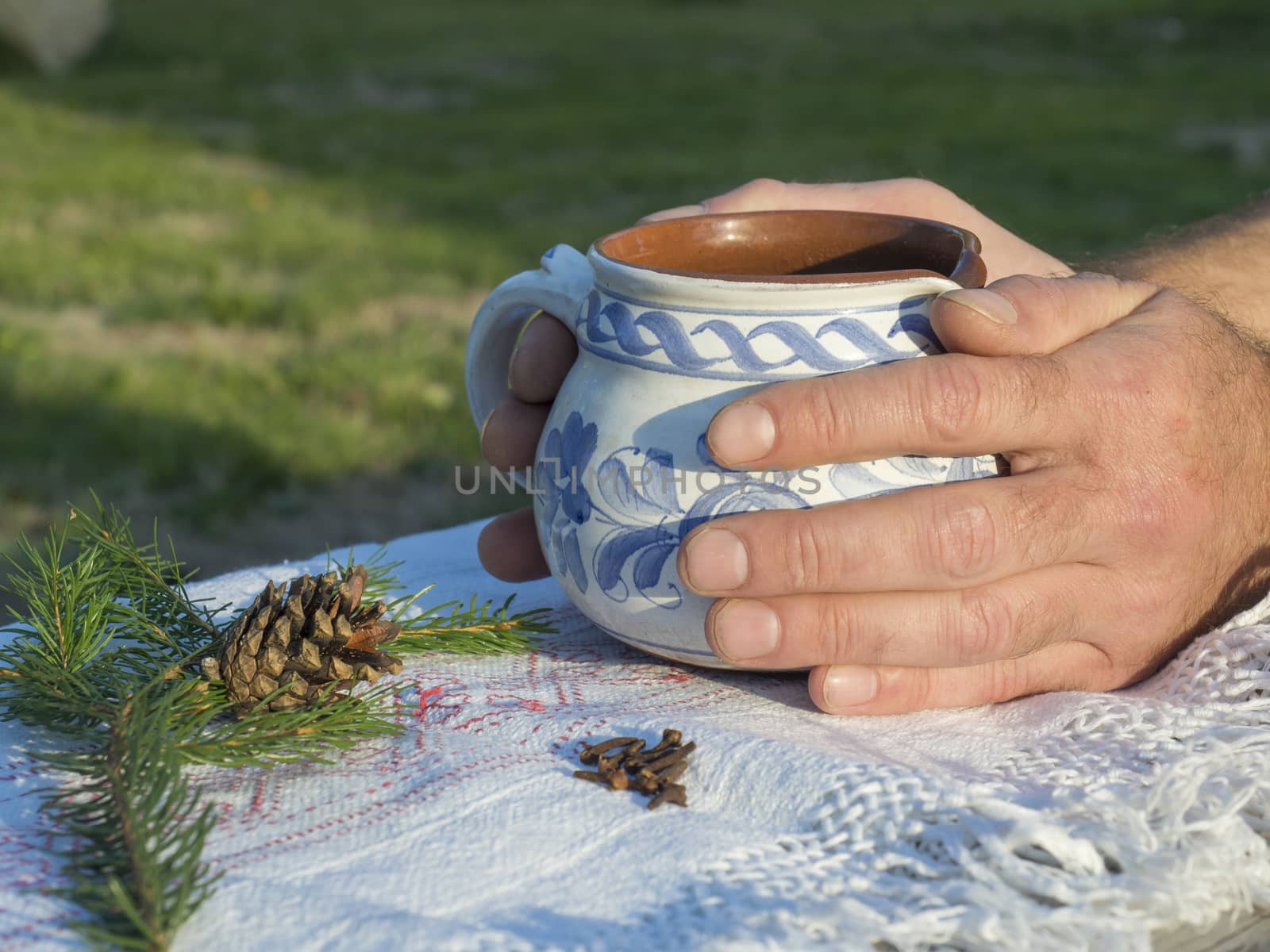 Close up hand of male holding hot smoking tea in rustic white blue painted ceramic cup, white tablecloth, green pine branches, cone and cloves spices, green background, golden light. Winter time coming concept by Henkeova