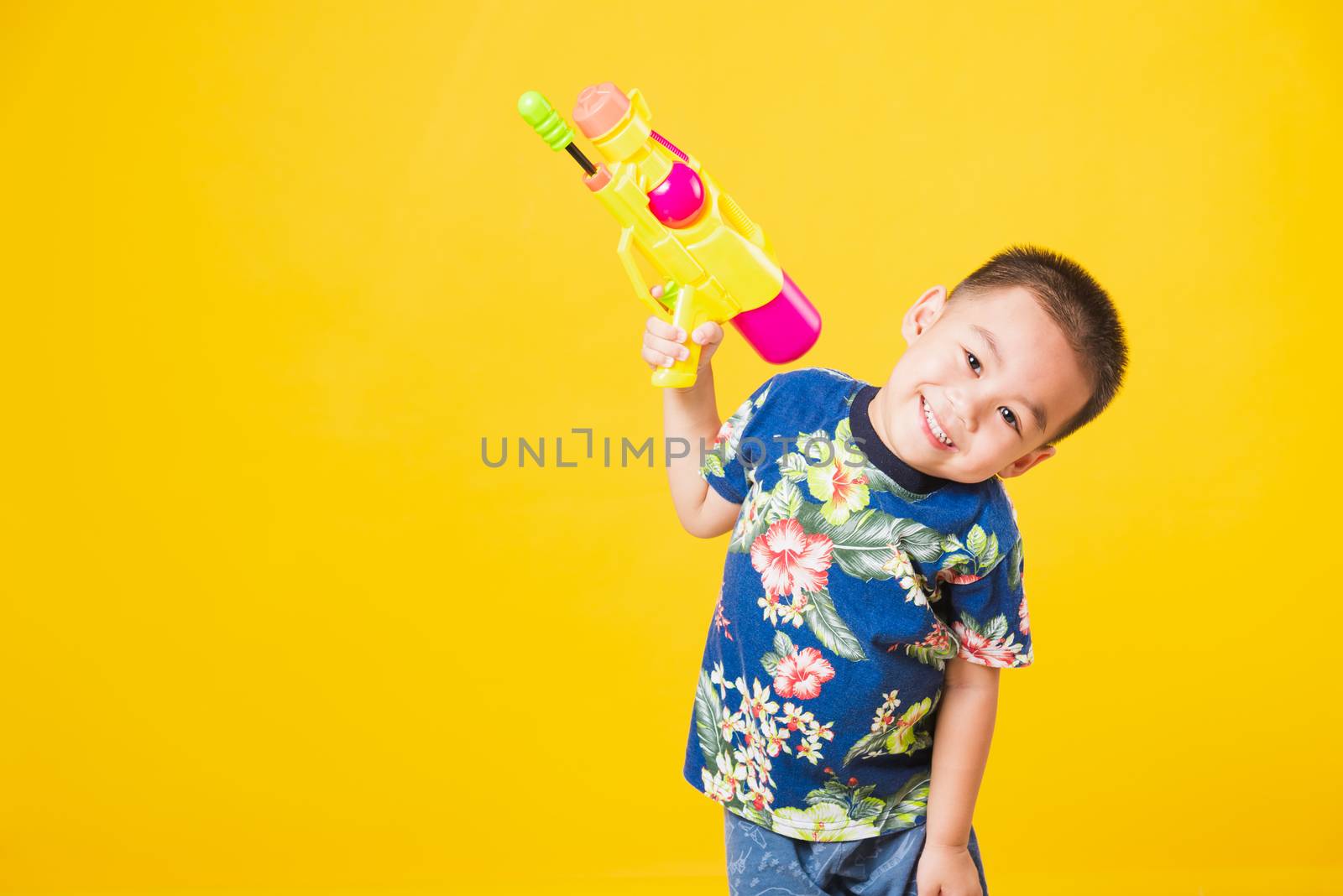 Portrait happy Asian cute little children boy smile standing so happy wearing flower shirt in Songkran festival day holding water gun, studio shot on yellow background with copy space