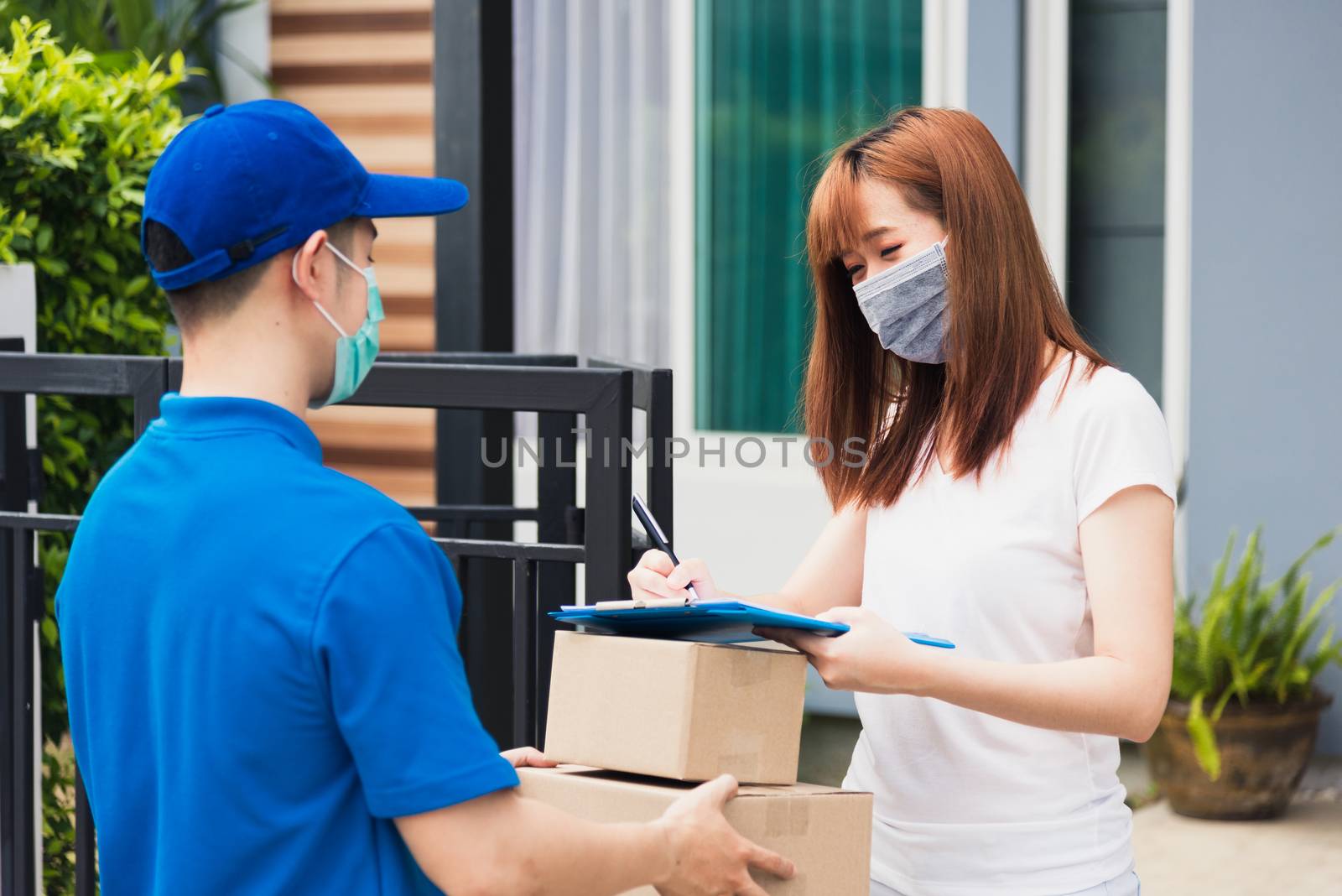 Asian young delivery man with package post boxed he protective face mask service woman customer using pen signature on paper book at front home, under curfew quarantine pandemic coronavirus COVID-19