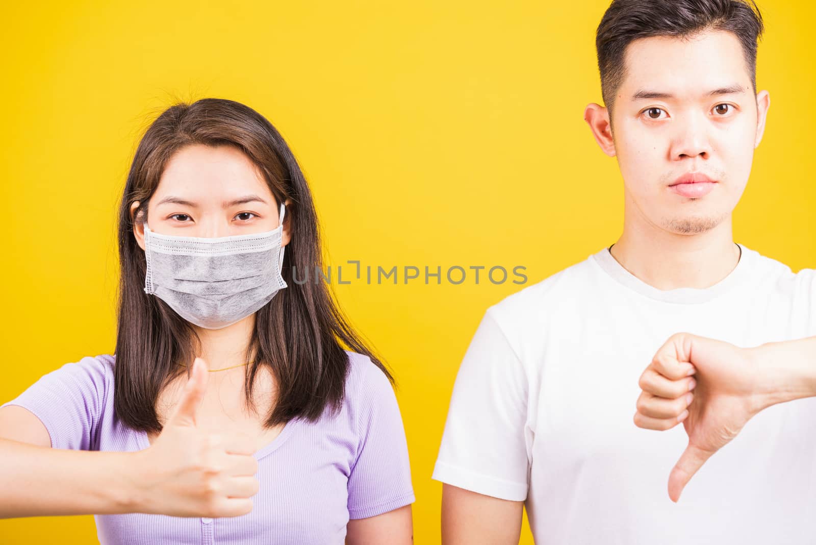 Asian beautiful woman and handsome man wearing protection face mask against coronavirus, show finger thumb UP and DOWN, studio shot isolated on yellow background, COVID-19 or corona virus concept