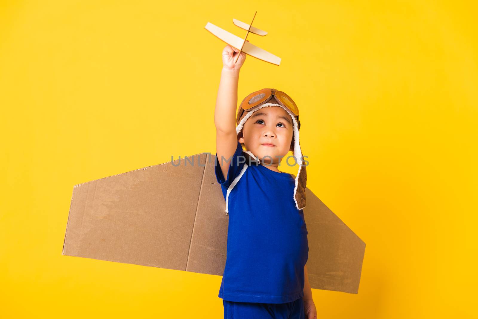 Happy Asian handsome funny child or kid little boy smile wear pilot hat play and goggles with toy cardboard airplane wings fly hold plane toy, studio shot isolated yellow background, Startup freedom