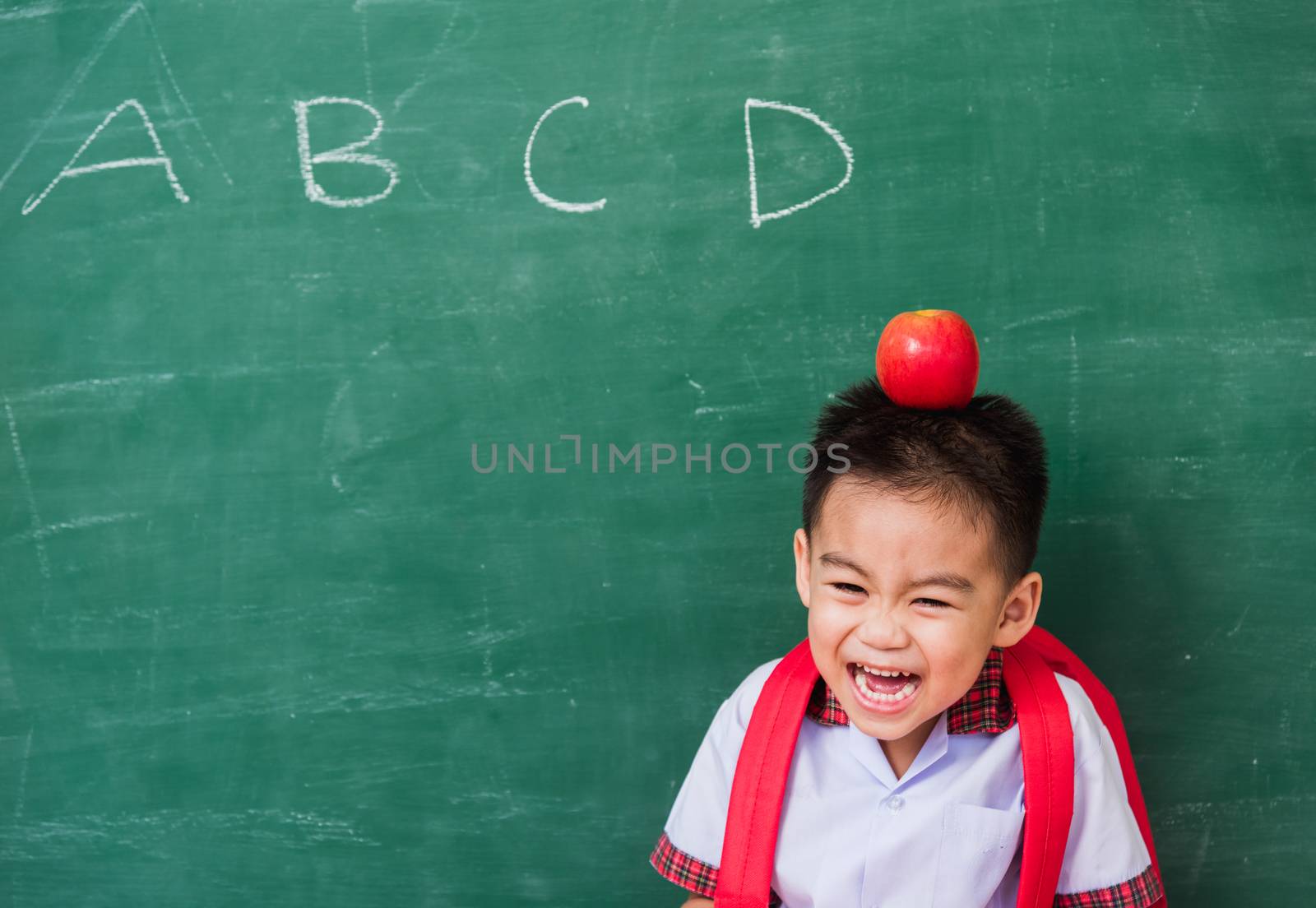 Back to School. Happy Asian funny cute little child boy from kindergarten in student uniform with school bag and red apple on head smiling on green school blackboard, First time to school education