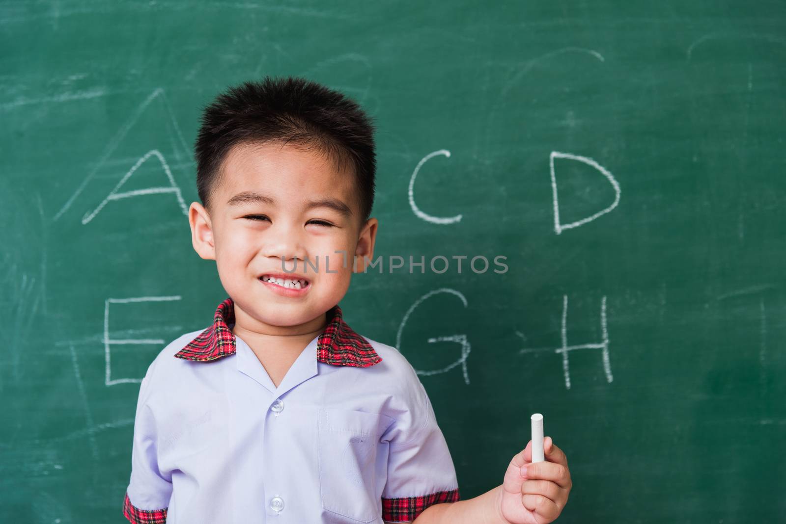 Back to School. Happy Asian funny cute little child boy kindergarten preschool smile in student uniform hold white chalk after write ABC with on green school blackboard, First time to school education