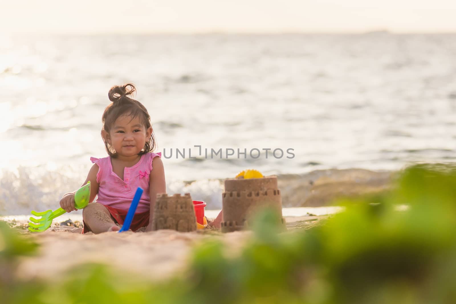 Happy fun Asian child cute little girl playing sand with toy sand tools at a tropical sea beach in holiday summer on sunset time, tourist trip concept