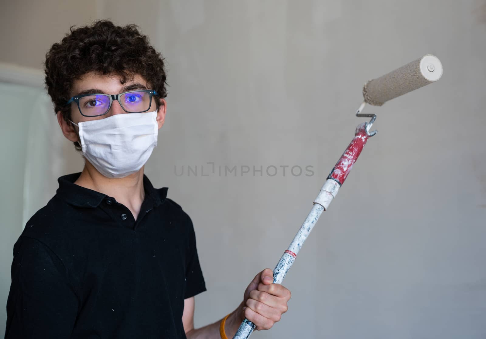 Renovate the walls of the house by painting them in the time of the coronavirus: the Caucasian boy has a mask to protect himself from the virus. Pass the impregnated roller of gray color on the wall.