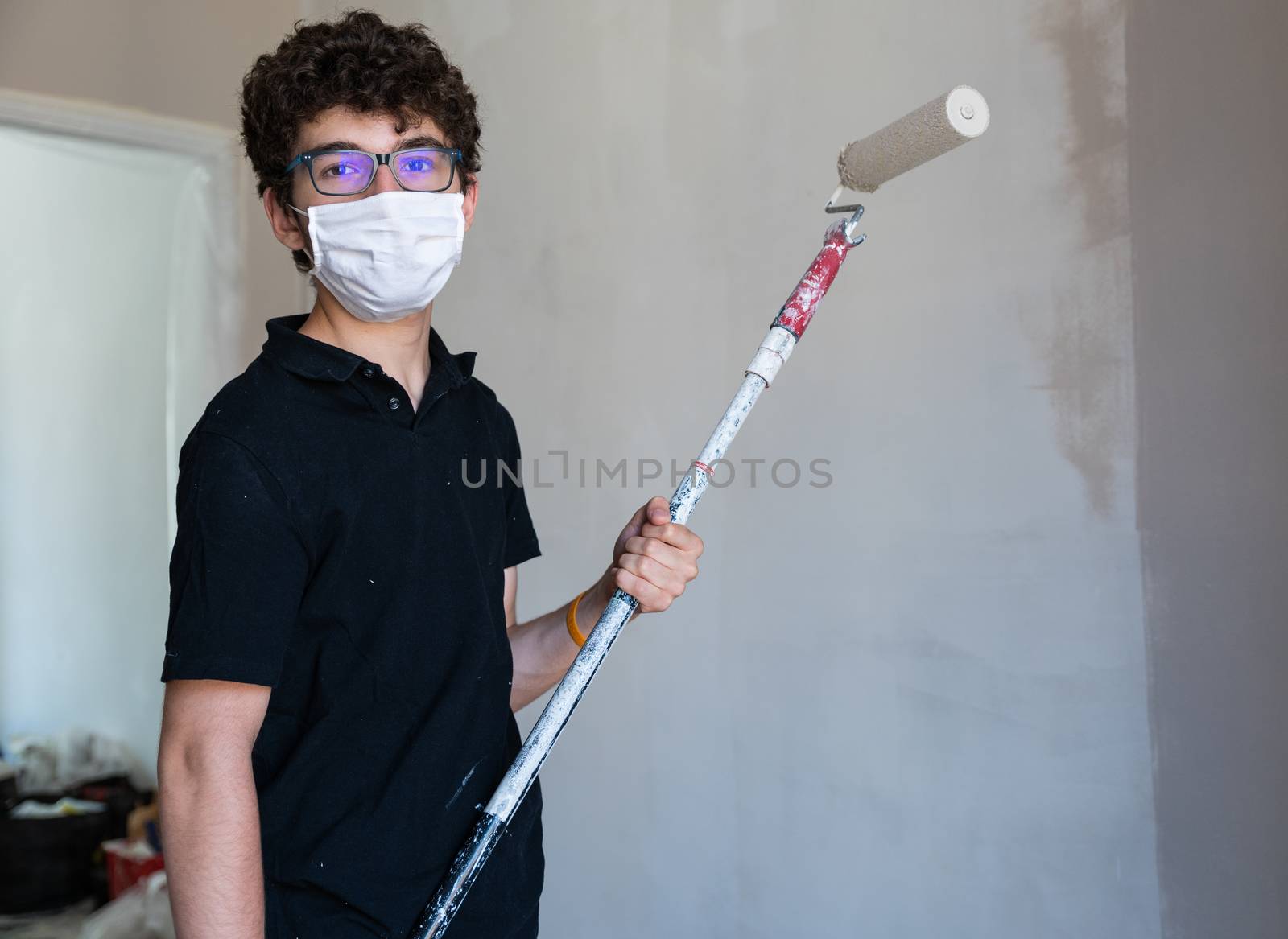 Renovate the walls of the house by painting them in the time of the coronavirus: the Caucasian boy has a mask to protect himself from the virus. Pass the impregnated roller of gray color on the wall.