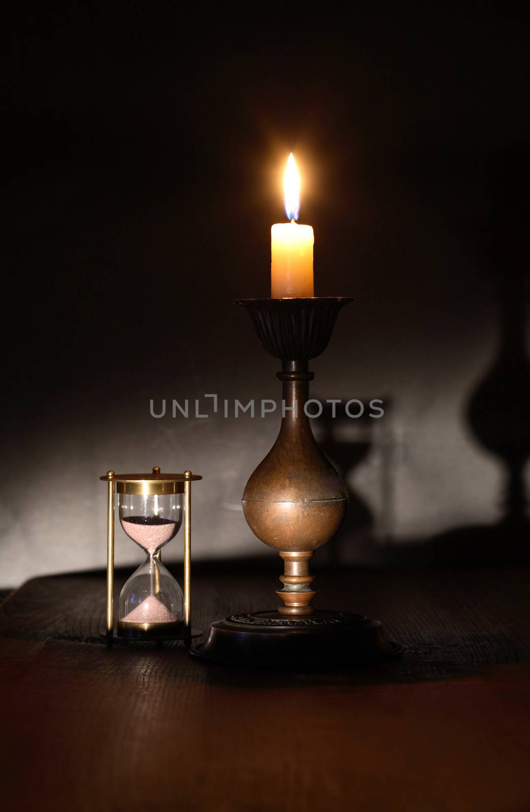 Elegant brass candlestick with lighting candle near hourglass on dark background