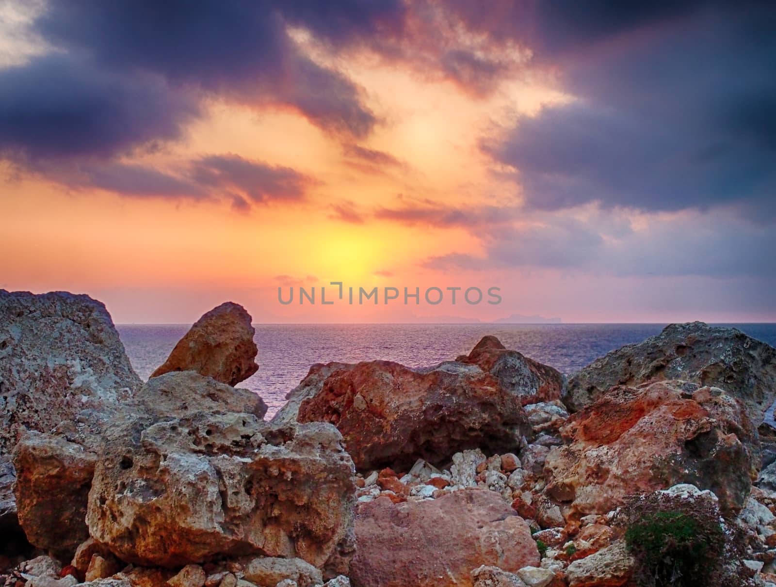 a glowing golden sunset surrounded by illuminated clouds over a calm Mediterranean sea with a foreground of coastal rocks colored by the evening light