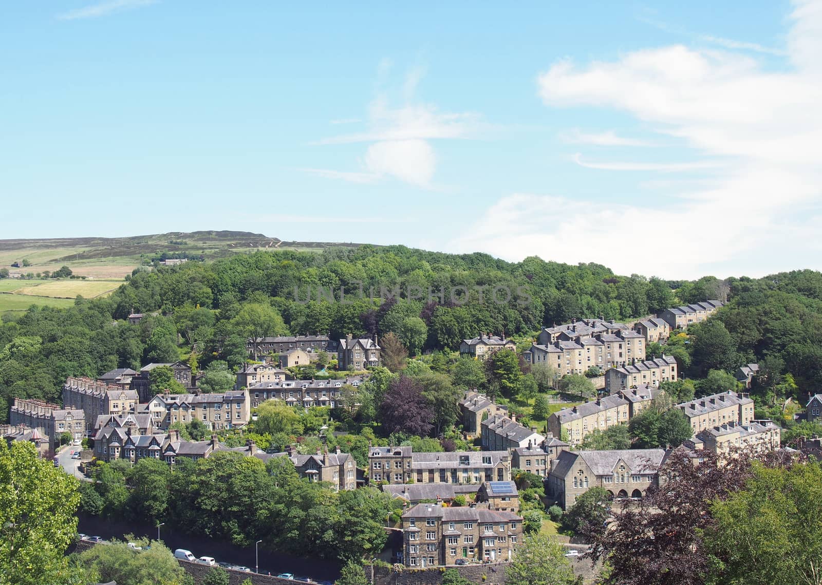 a scenic aerial view of the town of hebden bridge in west yorkshire with streets of stone houses and roads between trees and a blue summer cloudy sky