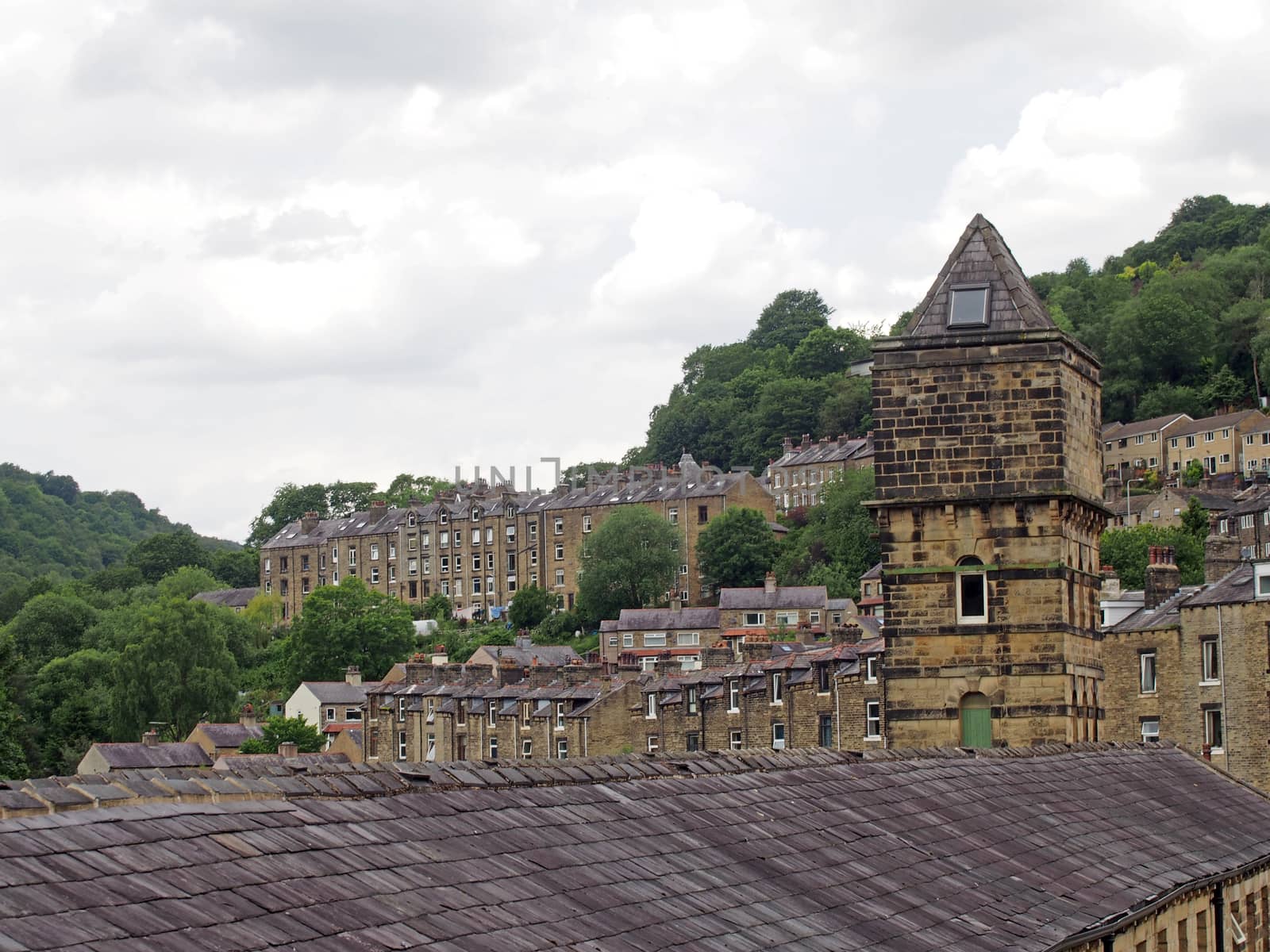 a view of the steep hillside streets in hebden bridge between summer trees with the tower of the historic nutclough mill building at the front