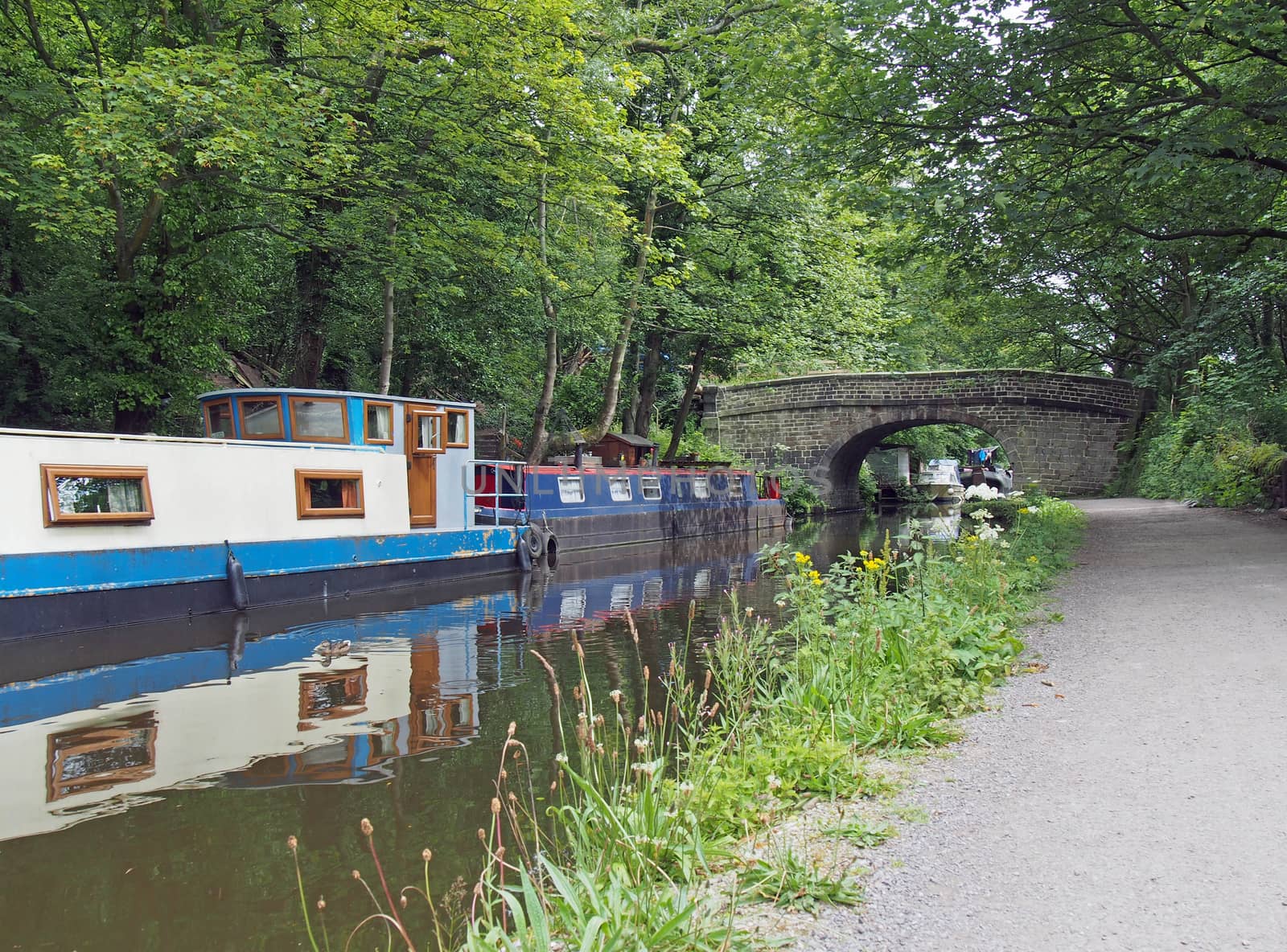 narrow boats and barges moored on the rochdale canal in hebden bridge bext to an old stone footbridge surrounded by green summer trees