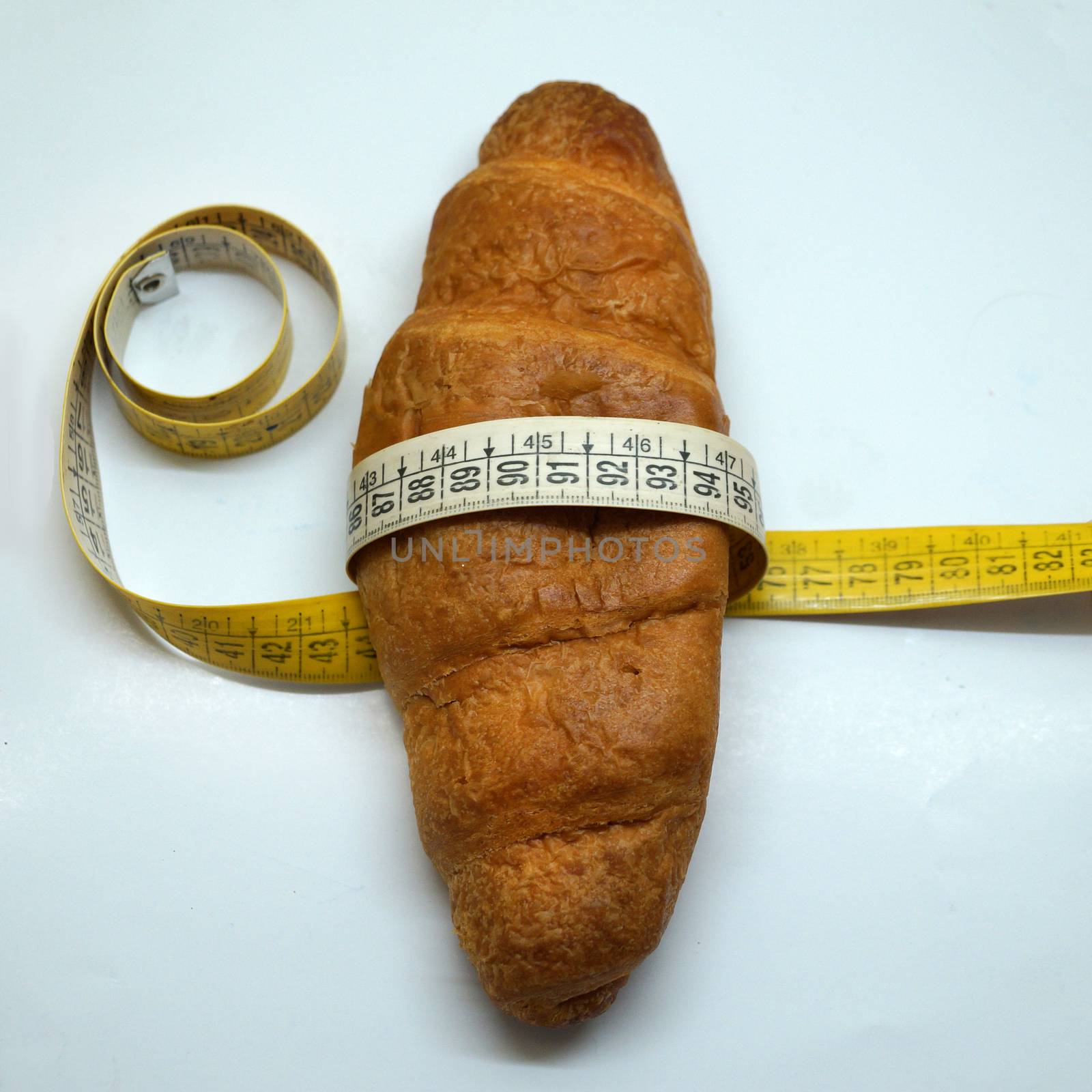 soft measuring ruler wrapped around a croissant as a symbol of unhealthy nutrition by Annado