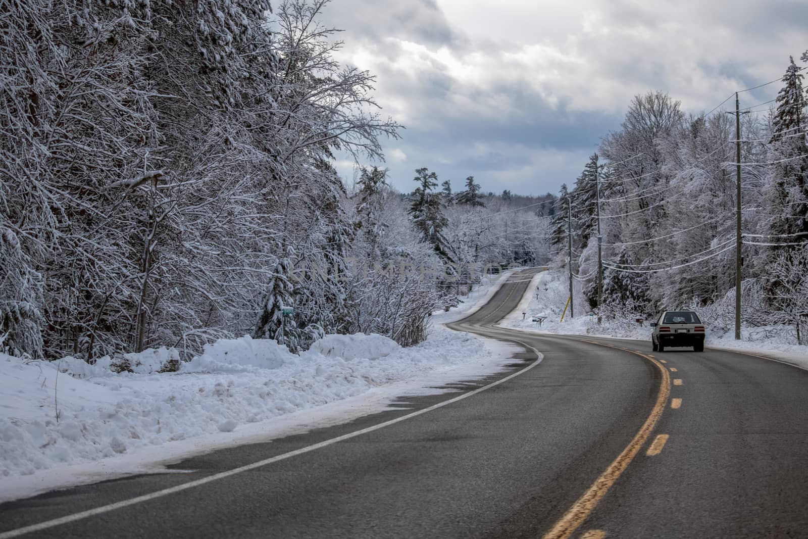 An older car is driving down a winding country road in the winter, with forest trees on either side of the road coated in a thick layer of fresh snow.