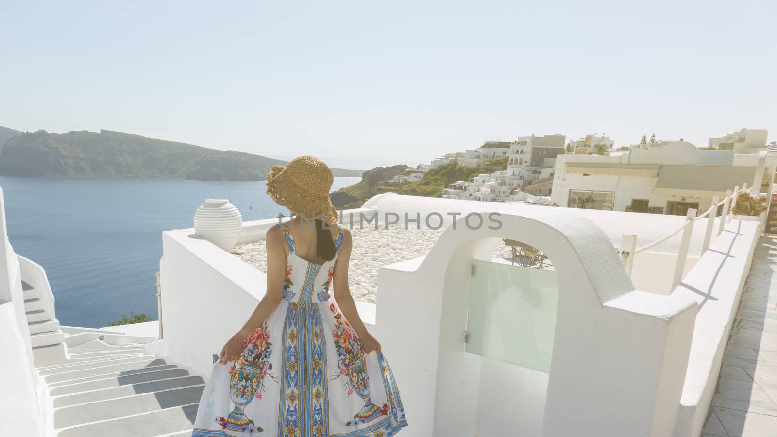 Back side of woman tourist walking on stairs in Oia village, Santorini with the mediterranean sea. Santorini Europe summer destination.