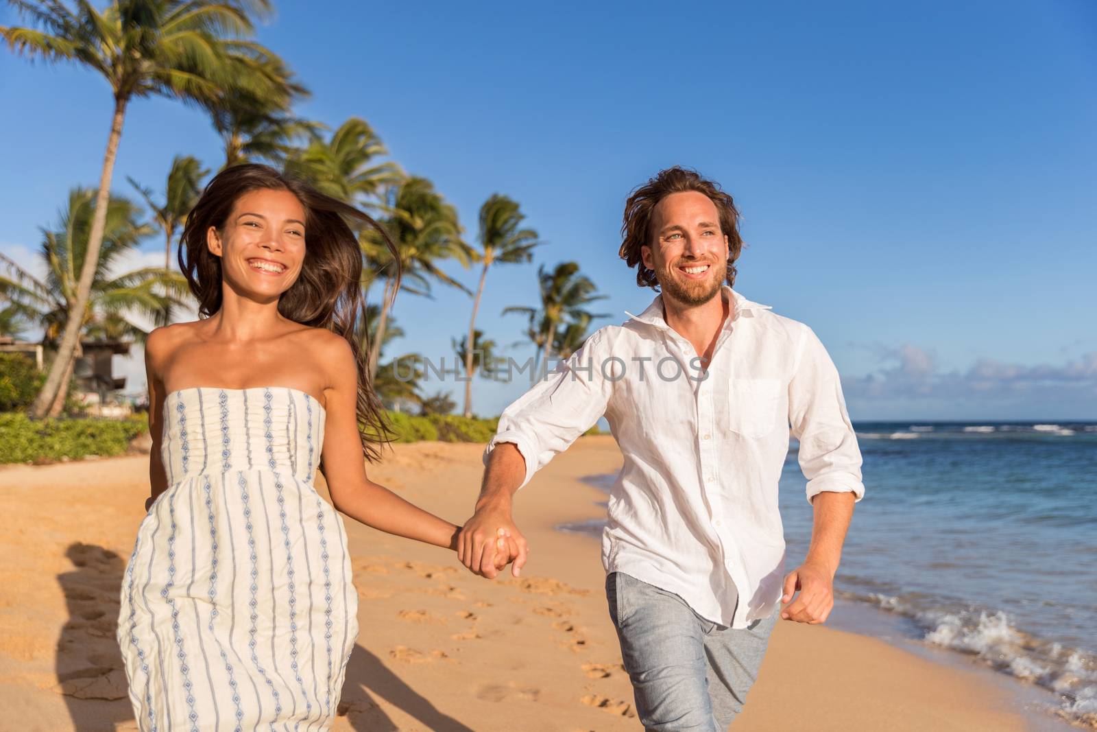 Happy newlyweds couple running on the beach holding hands, honeymoon travel. Asian caucasian multiracial people having fun together.