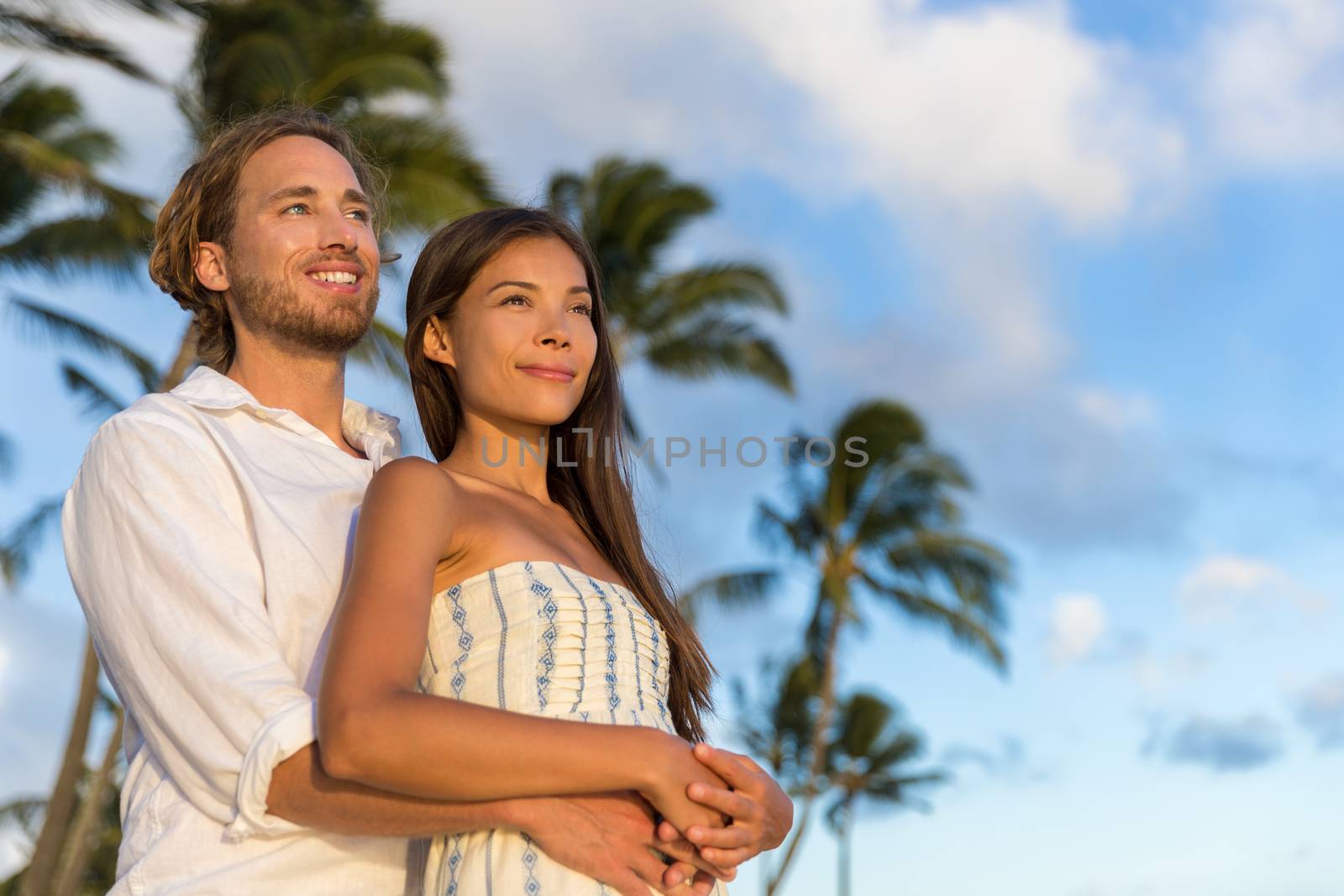 Relaxing couple watching sunset on summer travel vacations. multiracial people enjoying tropical holiday together hugging in love by Maridav