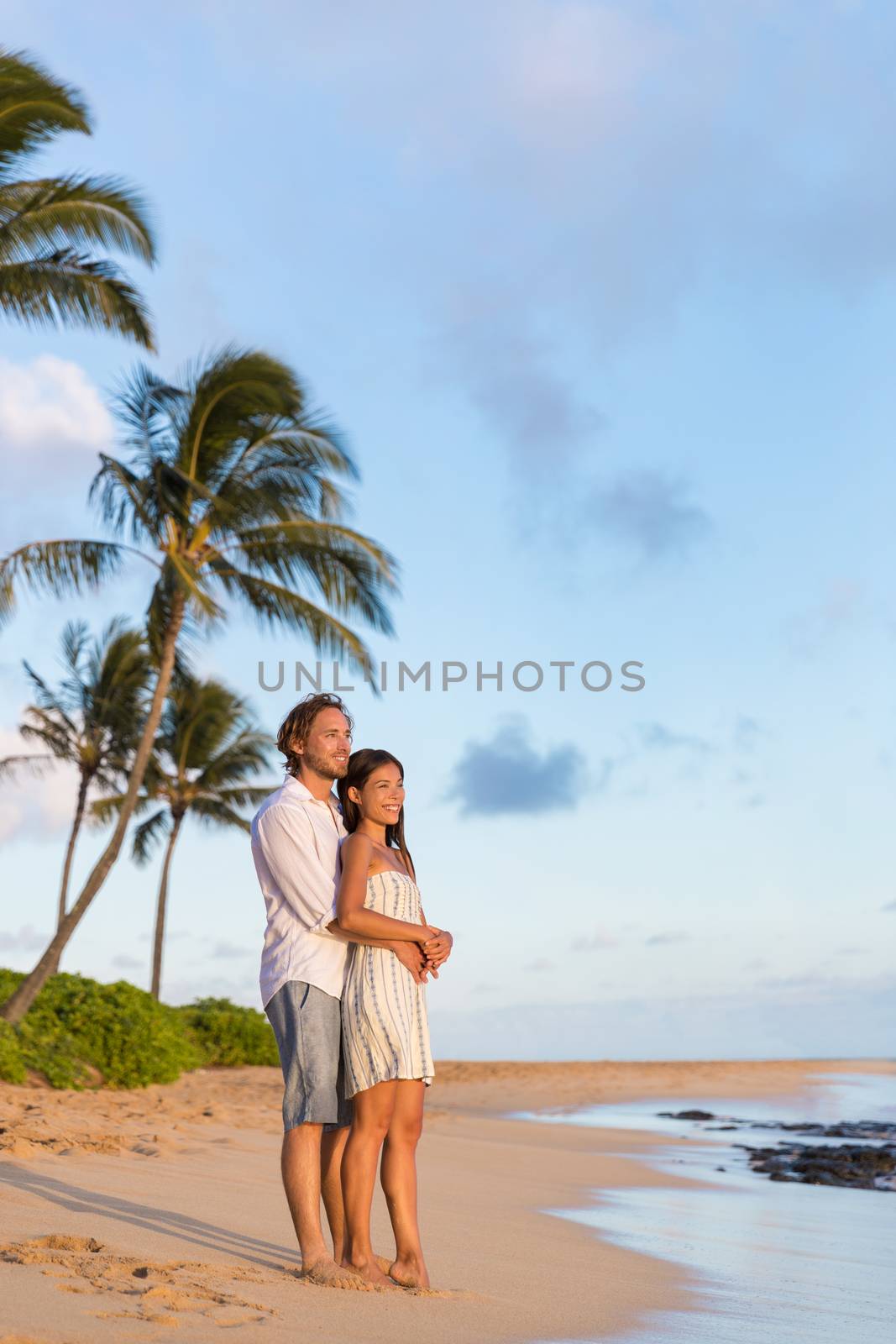 Relaxing couple watching sunset on beach vacation. multiracial people enjoying tropical holidays together hugging in love.