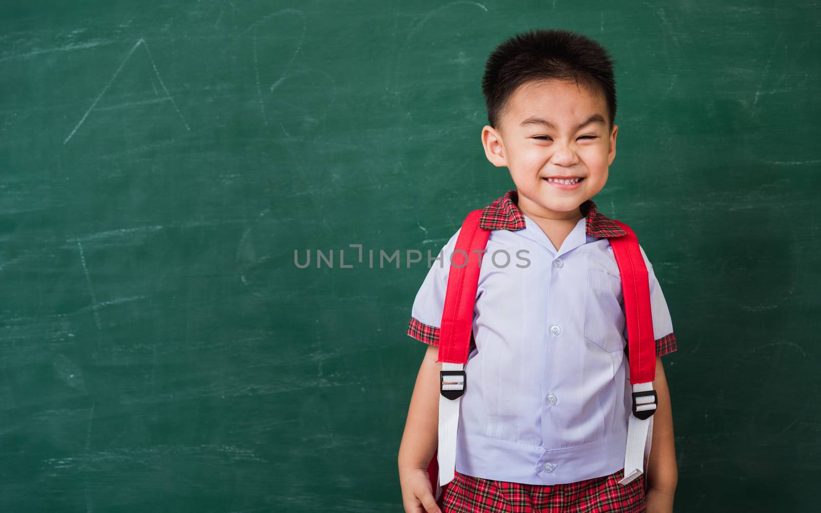 Back to School. Happy Asian funny cute little child boy from kindergarten in student uniform with school bag stand smiling on green school blackboard, First time to school education concept