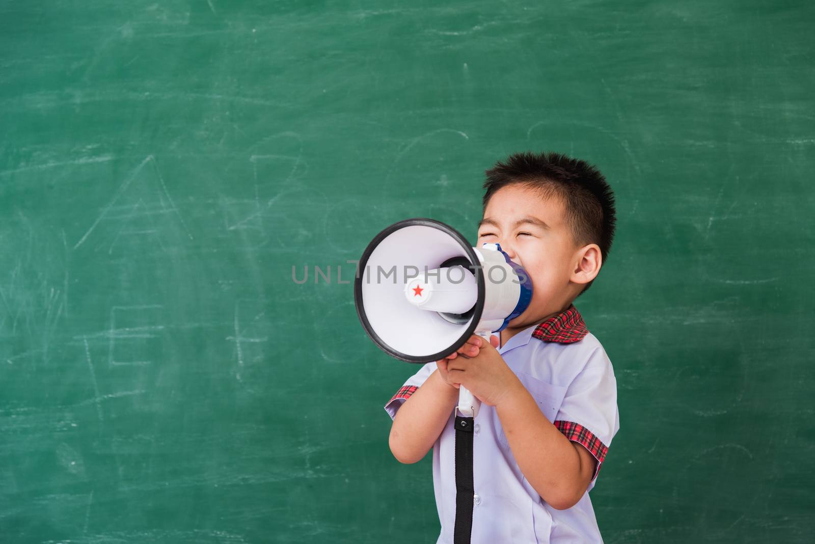 Back to School. Asian funny cute little child boy kindergarten preschool in student uniform speaking through megaphone against on green school blackboard, First time to school education concept