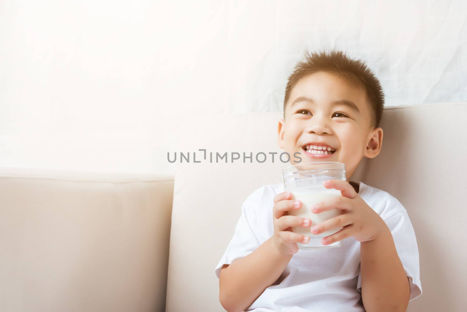 Close up of happy Asian little cute child boy hand holding milk glass he drinking white milk during sitting on the sofa at home after lunch. Daily life health care Medicine food