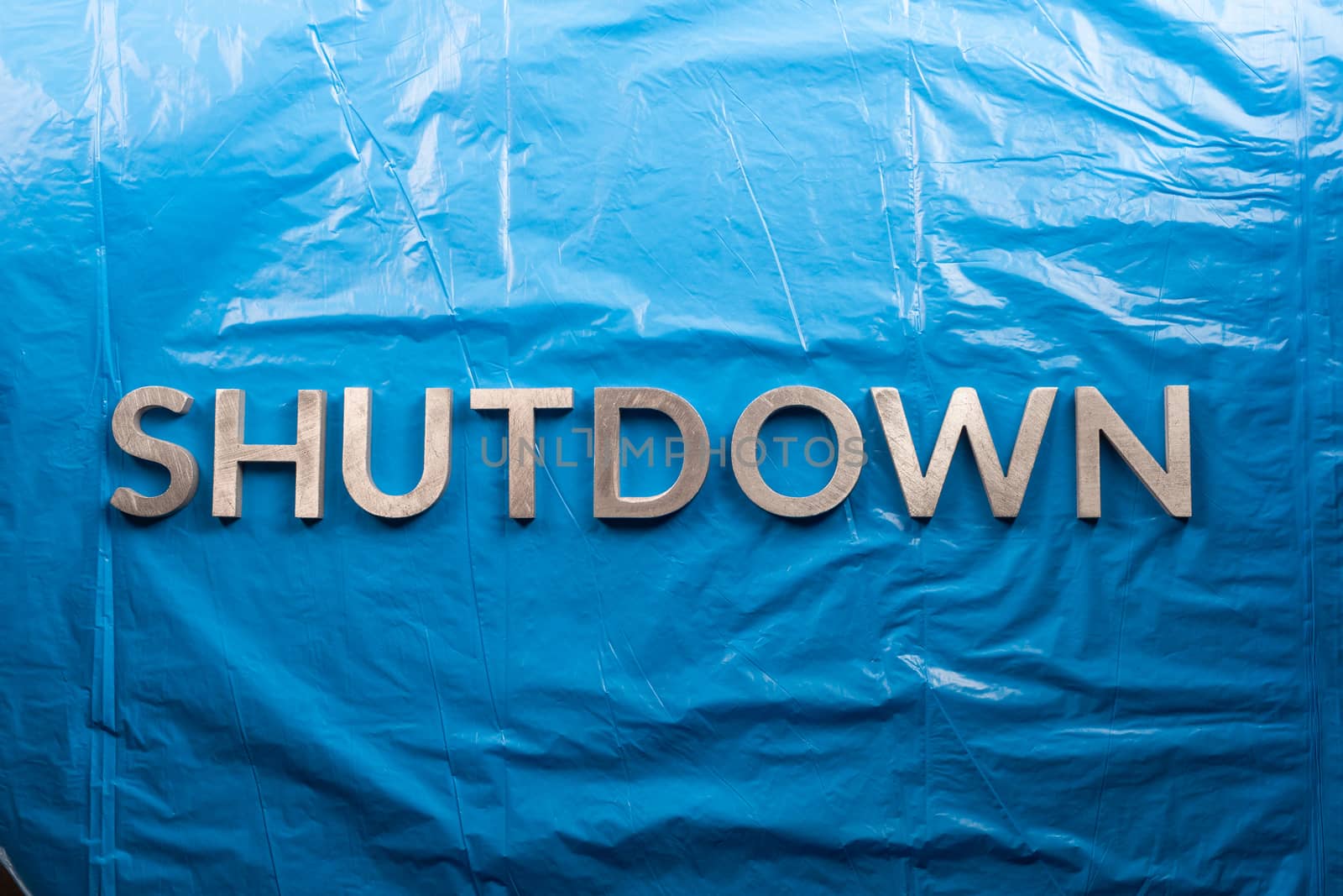 the word shutdown laid with silver metal letters on crumpled blue plastic film background in flat lay perspective.