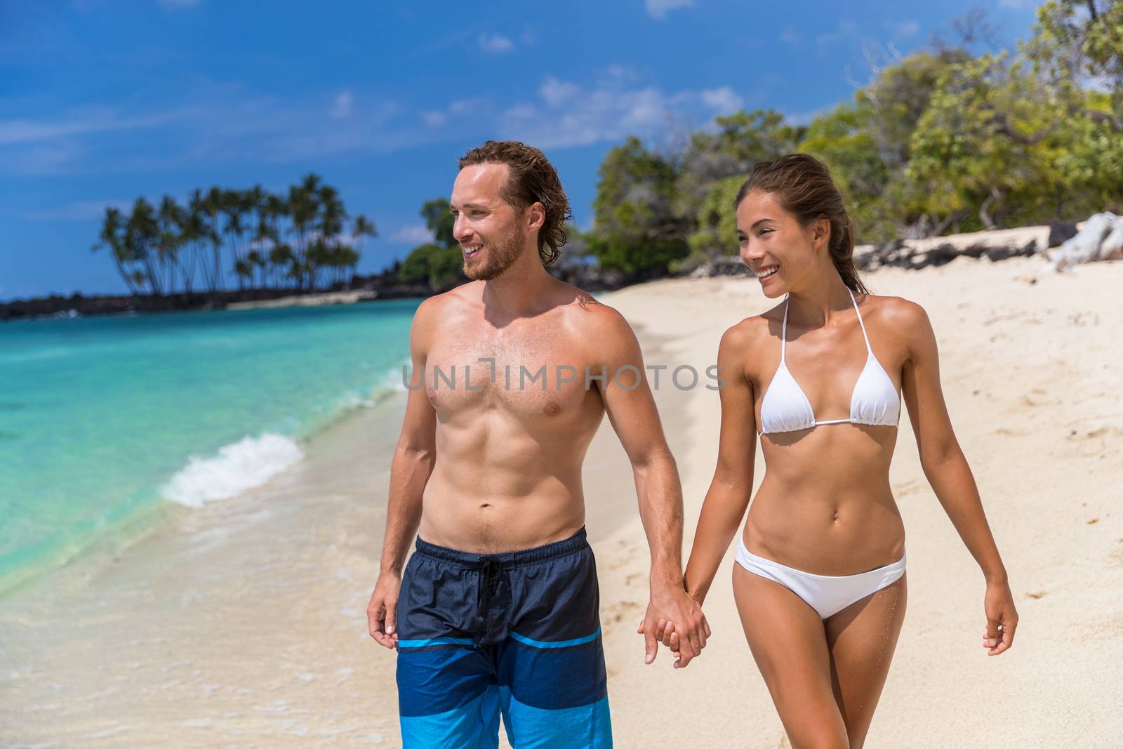 Beach bikini couple enjoying summer vacation travel holding hands on Hawaii tropical holidays. Sexy multiracial people with toned abs body by Maridav