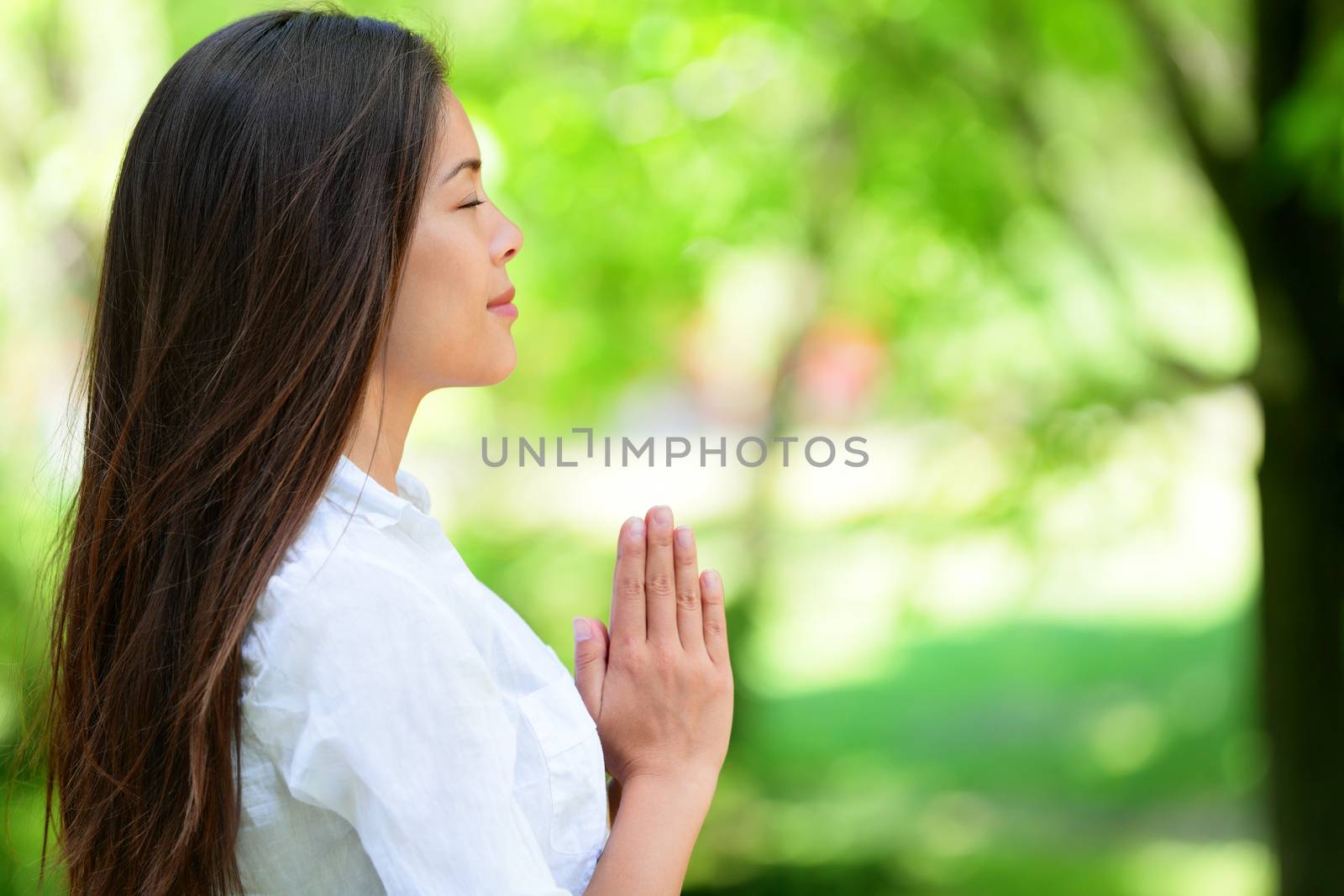 Relaxed young woman with hands clasped meditating in park. Profile view of woman in casuals. Attractive female with eyes closed practicing yoga in nature.
